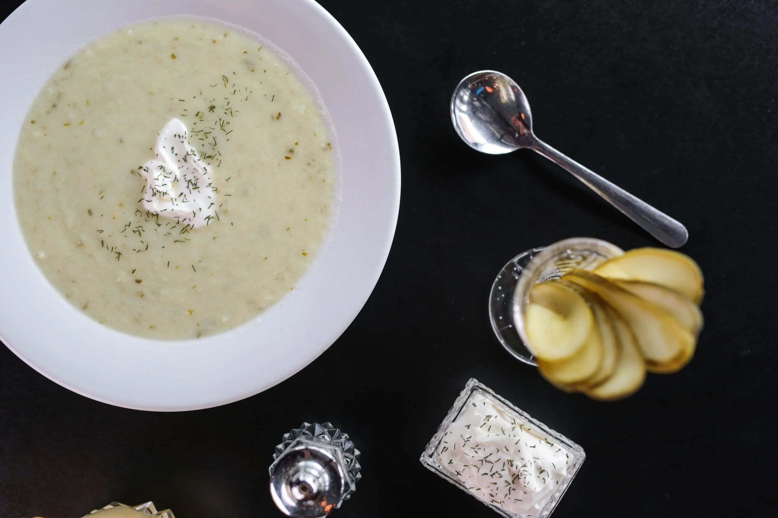 Photo of a bowl of pickle soup next to a container of salt, a soup spoon and a cup of pickles. Pickle soup is the best soup in st louis