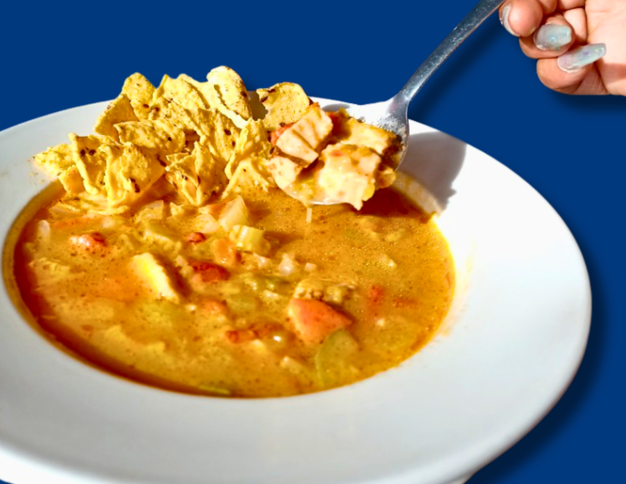 A white bowl of golden chicken tortilla soup with vegetables and shredded chicken, topped with crunchy tortilla chips; a spoon lifts a chip‑laden bite above the soup.