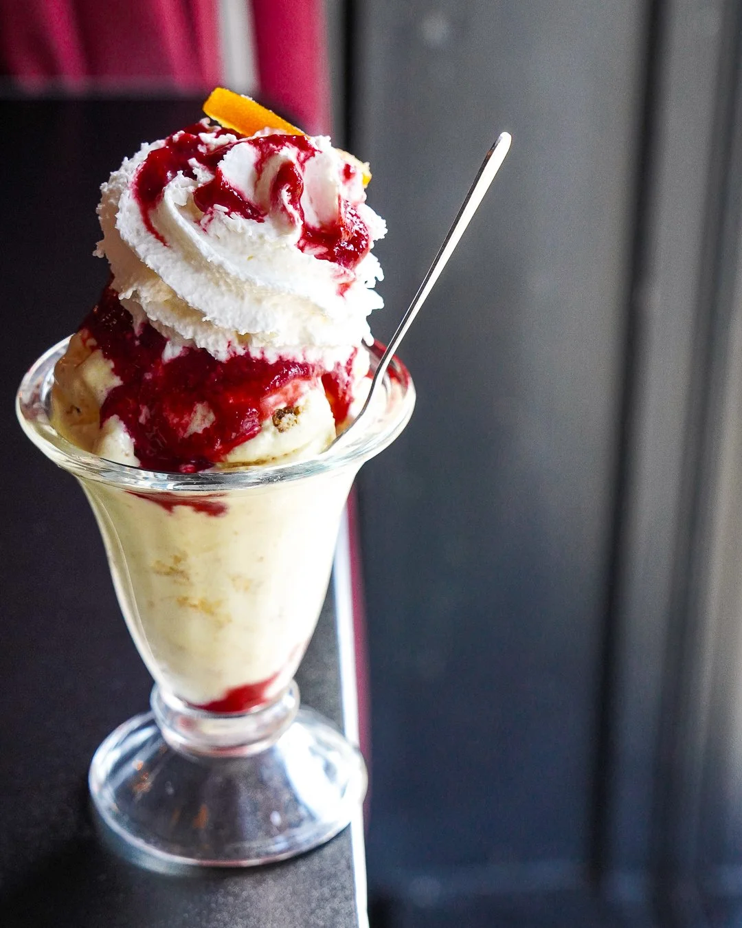 A glass sundae dish filled with Lemon Cheesecake ice cream, topped with raspberry sauce, whipped cream, and an orange peel garnish, with a spoon sticking out.
