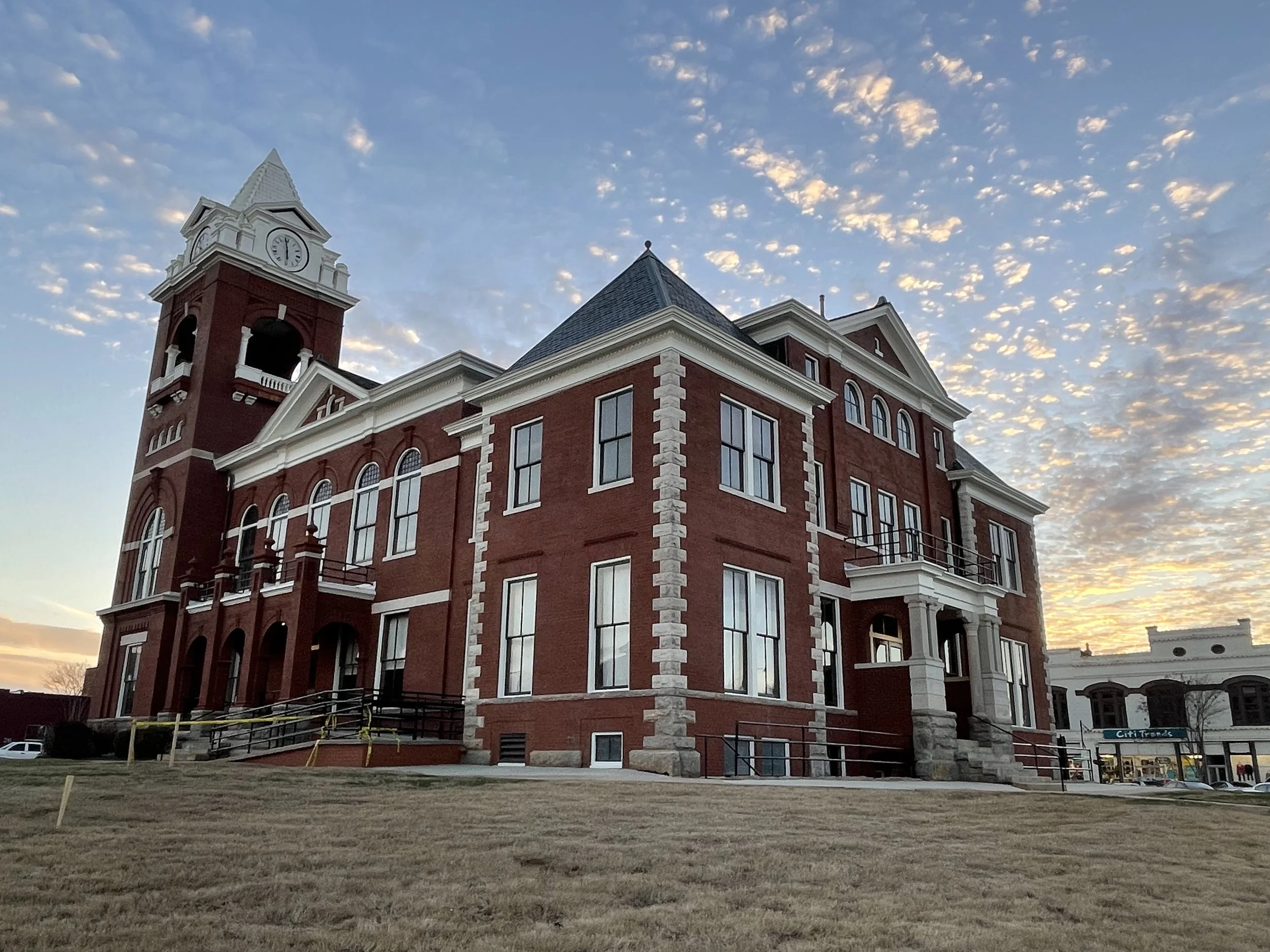 A red brick building with a clock tower and a pointed roof extends into a partly cloudy sky at sunset. The building has arched windows and a small balcony, with a grassy lawn and construction barriers in front.