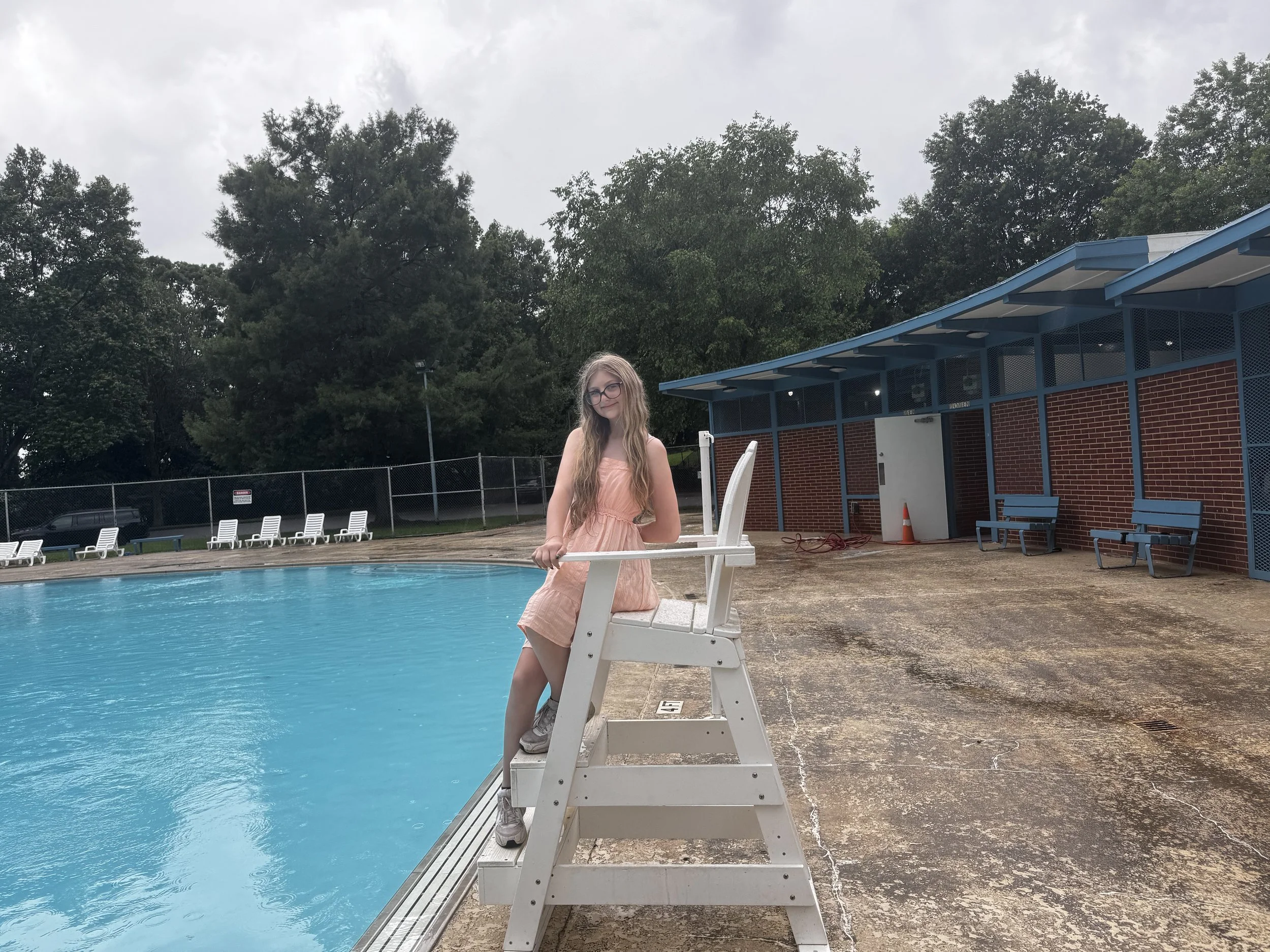 A young girl with long hair and glasses sitting on a lifeguard chair by a swimming pool, with trees and a building in the background.