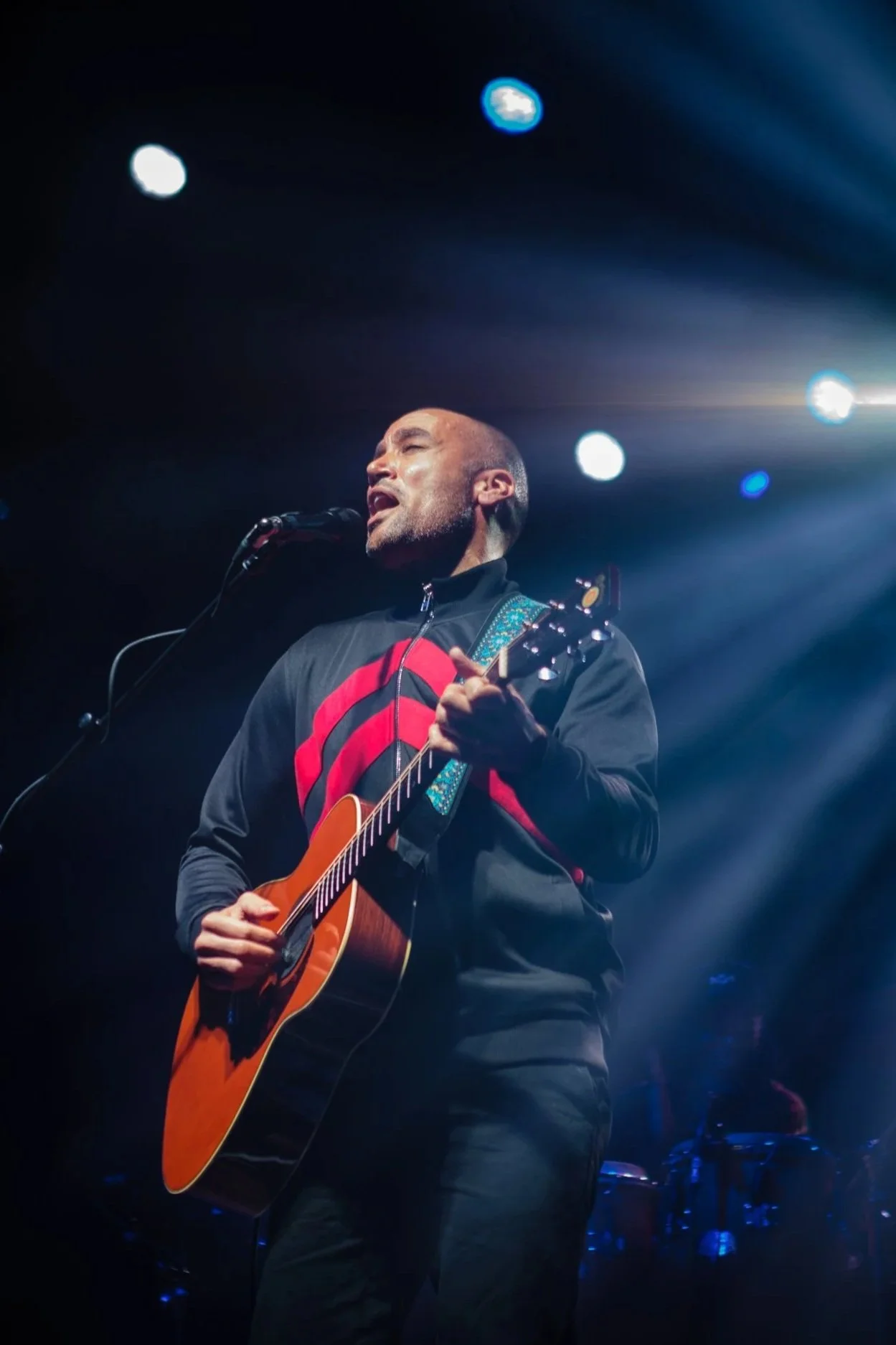 A man passionately singing and playing an acoustic guitar on stage, illuminated by bright stage lights and dark background.