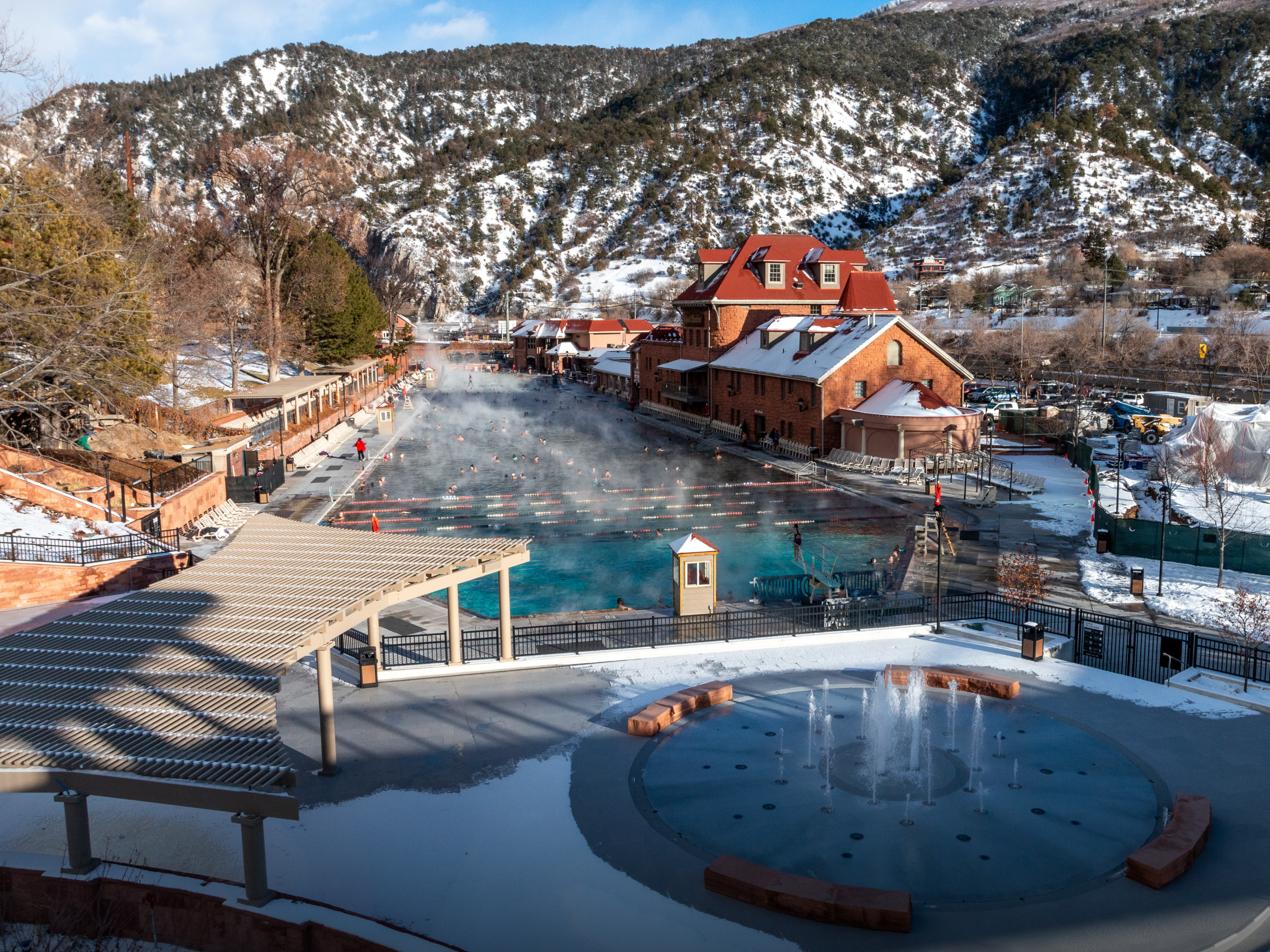 An outdoor hot spring or thermal bath facility in a snowy mountainous area, with steam rising from the water, surrounded by buildings and trees, and a mountain backdrop.