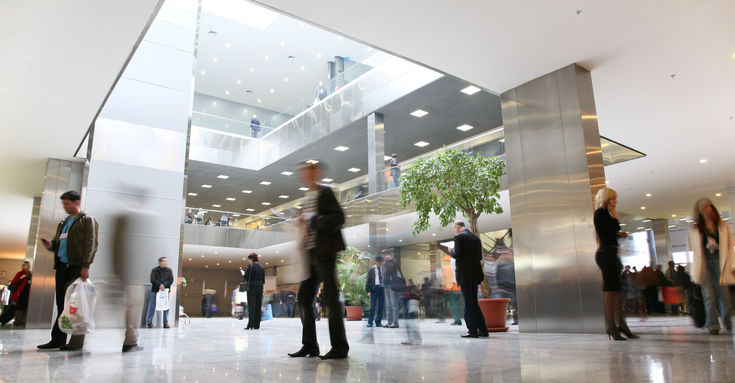 Commercial and Retail. Modern multi-story lobby with people walking and standing, some blurred due to motion, featuring a large indoor tree, shiny metallic walls, and a spacious open design with an upper level balcony.