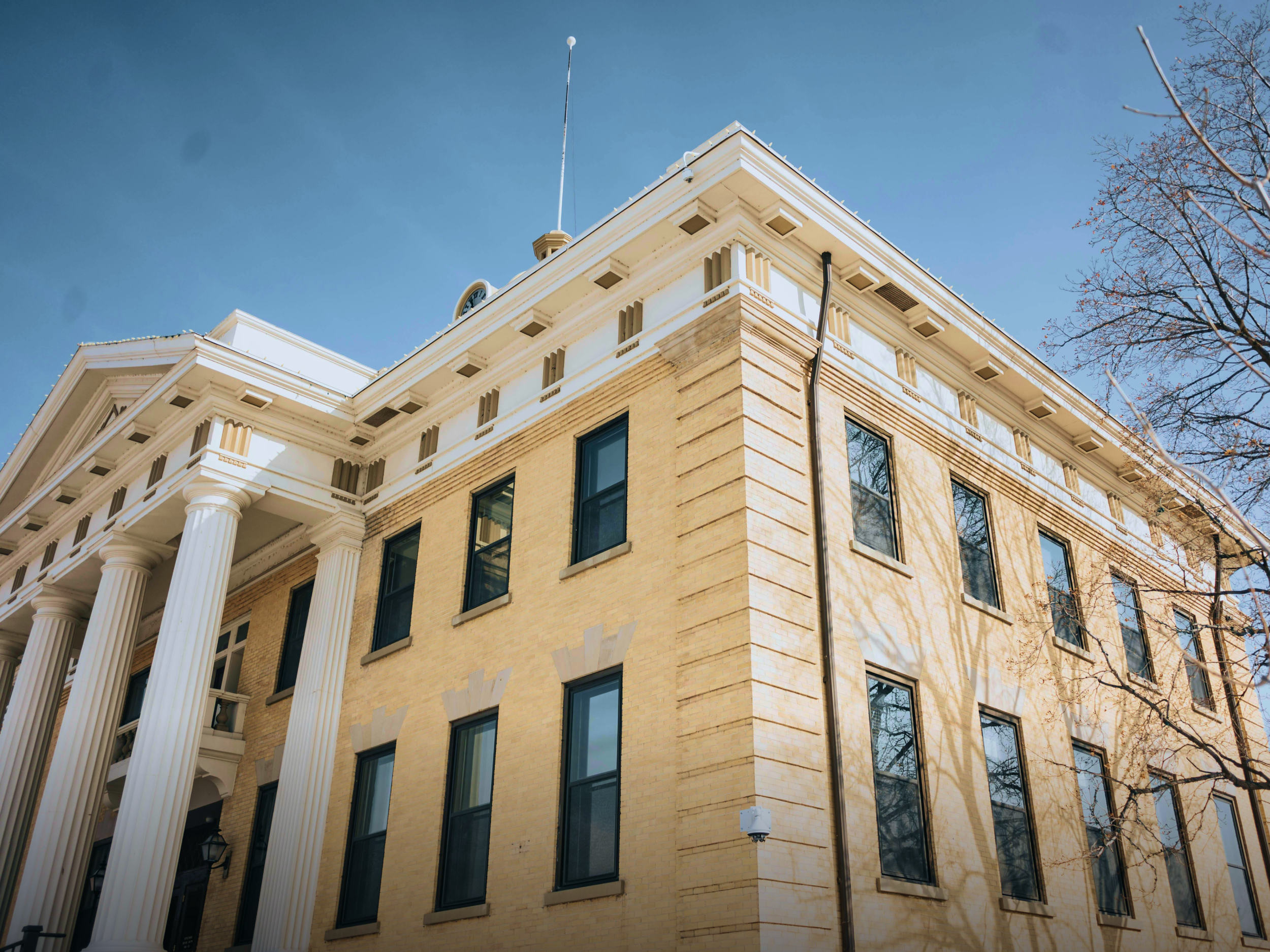 A historic building with classical architecture, featuring large white columns, ornate details, and multiple windows, set against a clear blue sky with leafless trees nearby.