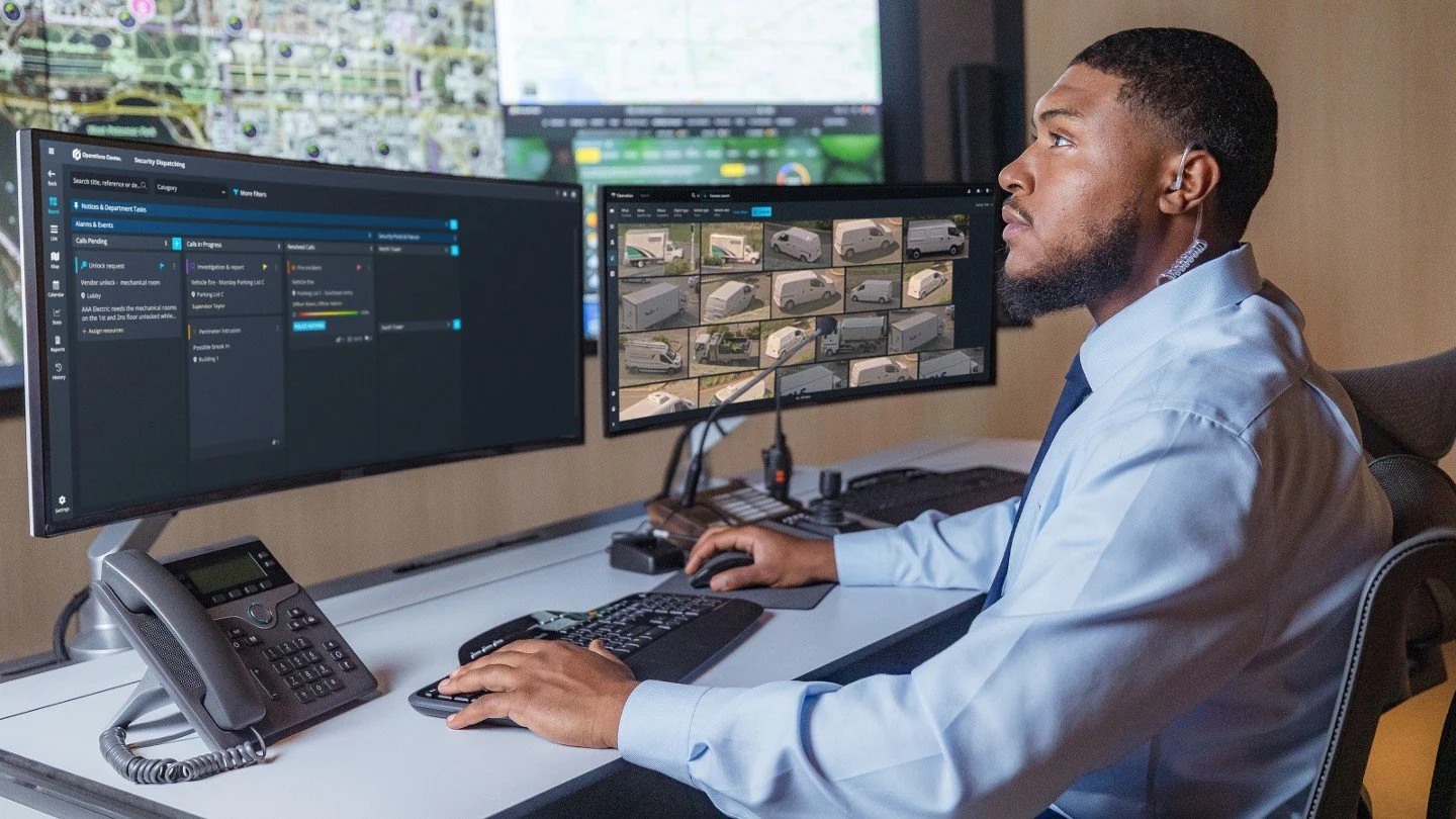 Genetec Security Operations. Man in office with multiple monitors displaying security camera footage and data monitoring screens.