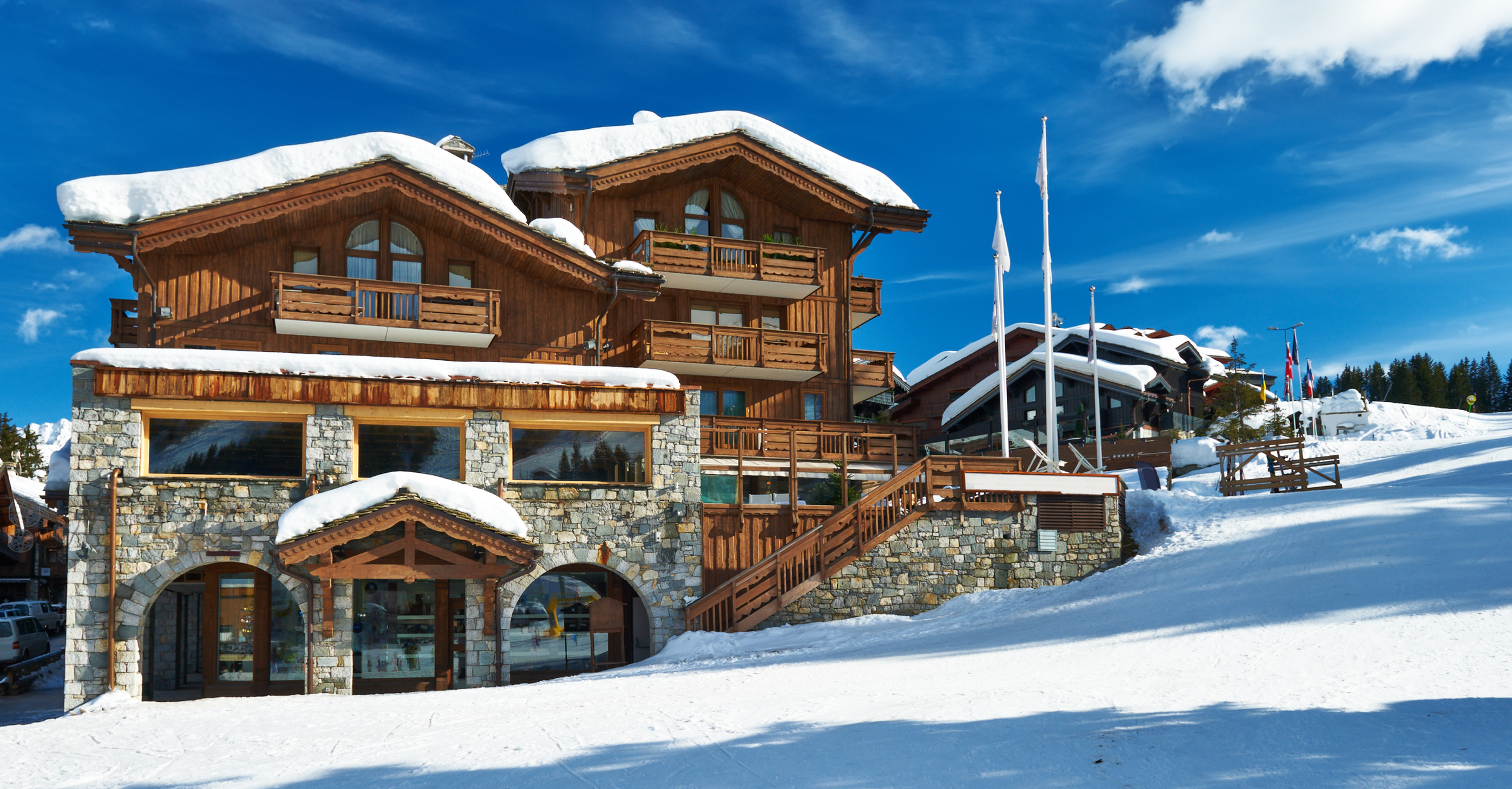 Hospitality. Large wooden and stone chalet covered in snow on a hillside under a bright blue sky with some clouds, with flags on poles and trees in the background.