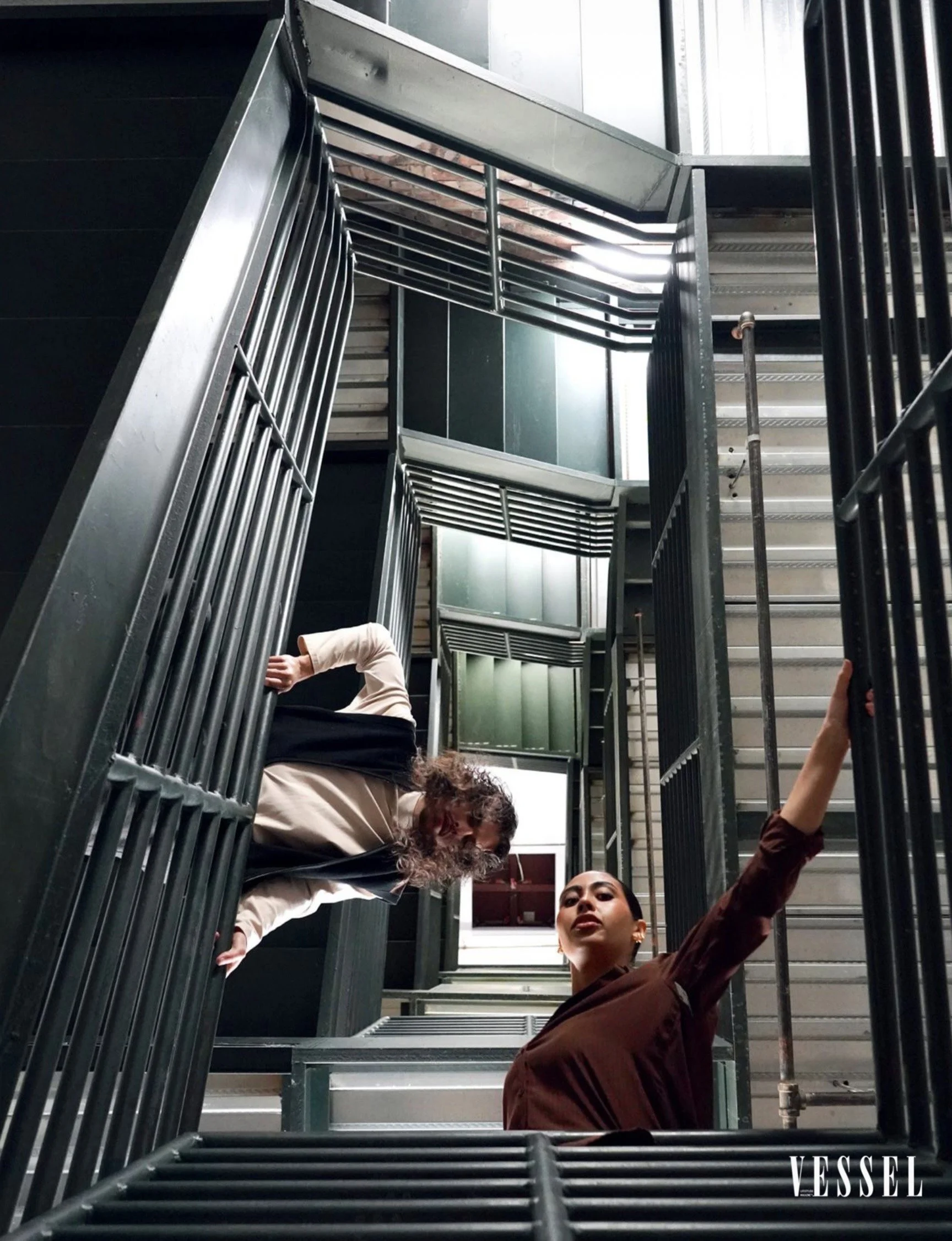 Multiple children and an adult on a spiral staircase, viewed from below, with metal railings and steps, in an indoor setting.