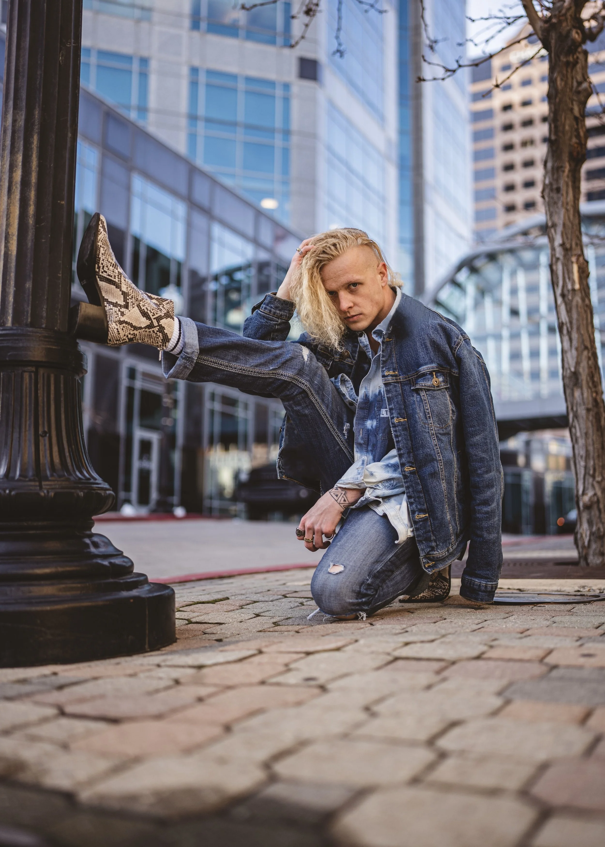 A person with blonde hair in a denim jacket and ripped jeans kneels on a brick sidewalk with one leg resting on a park lamp post, in an urban setting with tall glass buildings and a tree.