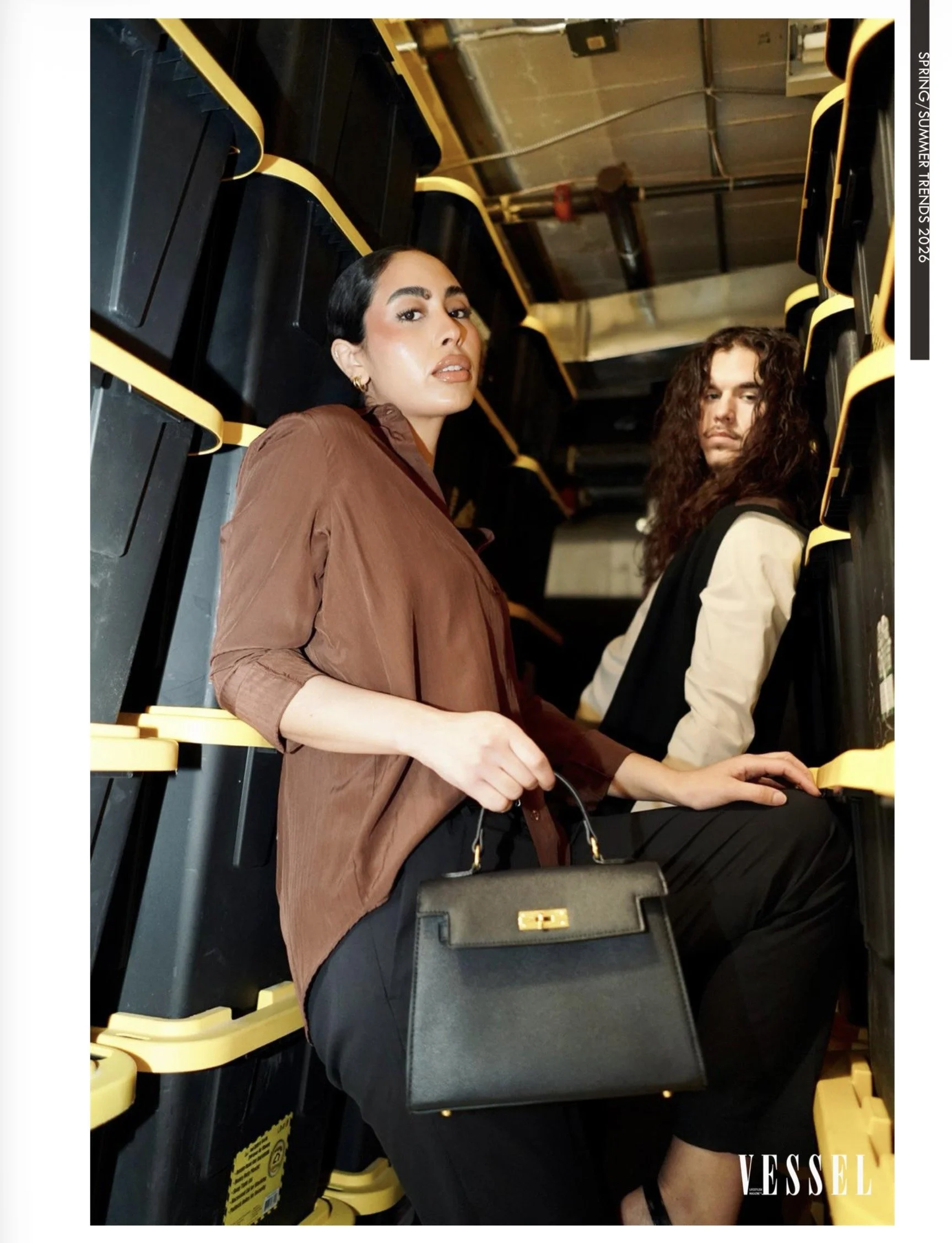 Two women sitting among yellow and black seats at a venue, with one woman wearing a brown jacket and holding a black handbag, and the other woman wearing a cream and black jacket with curly hair, both looking at the camera.
