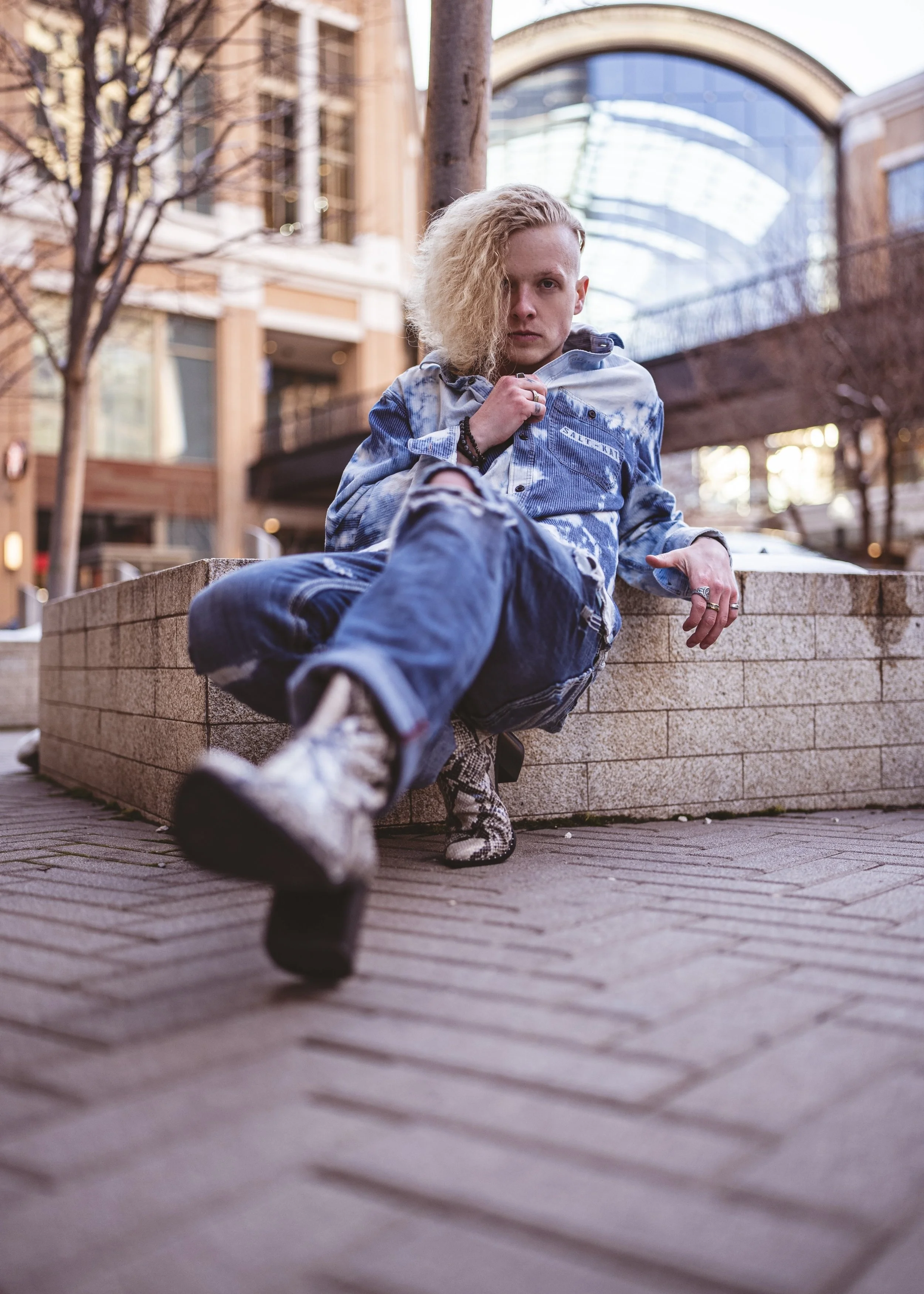 A young person with blond, curly hair sitting on a brick ledge in an urban area, wearing a ripped denim jacket and jeans, with a serious expression, one hand near their face and the other resting on the ledge. The background features modern buildings and leafless trees.