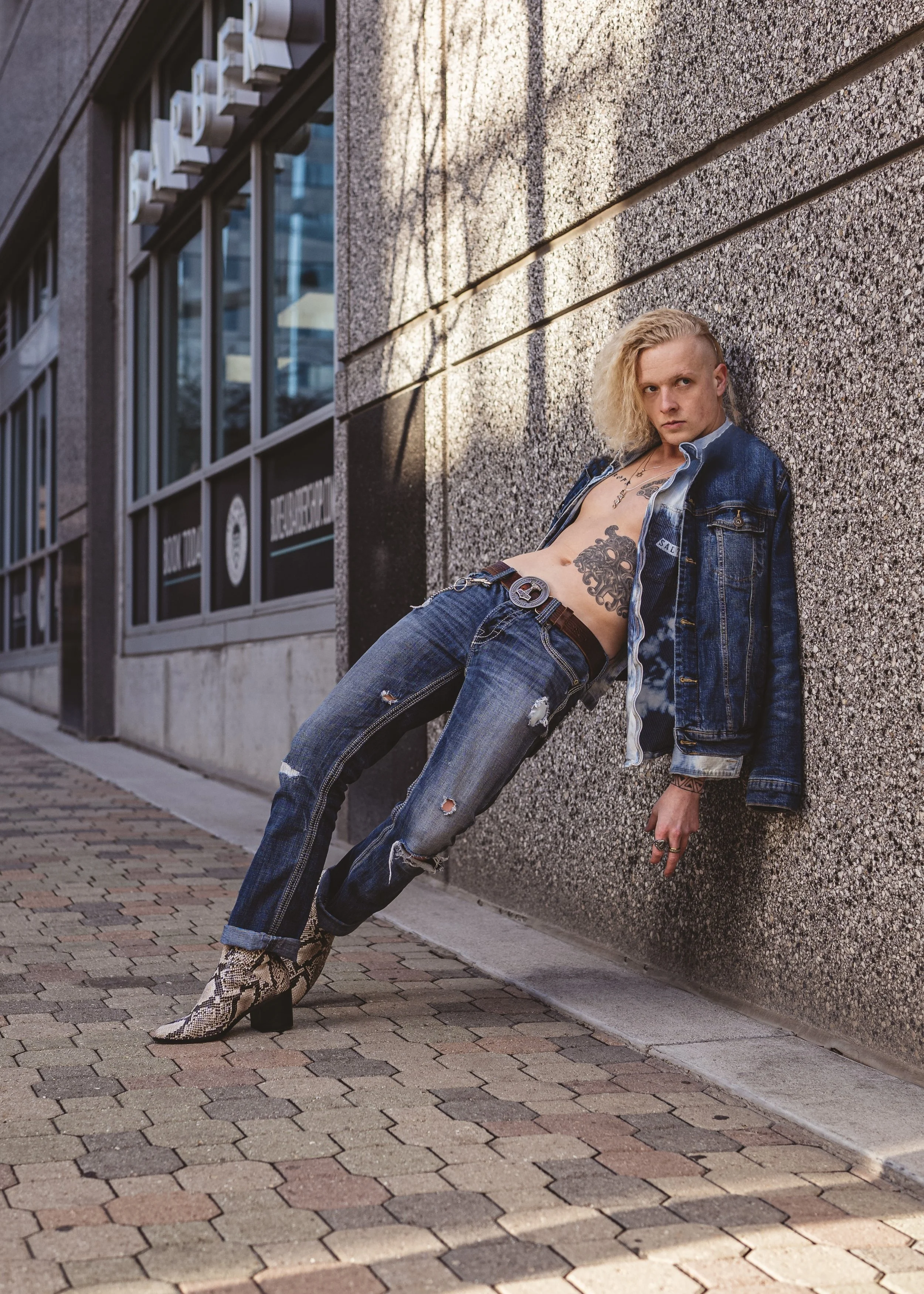 Fashionably dressed person with blonde curly hair leaning against a textured wall in an urban setting, wearing ripped jeans, snake print boots, and an open denim jacket, looking at the camera.