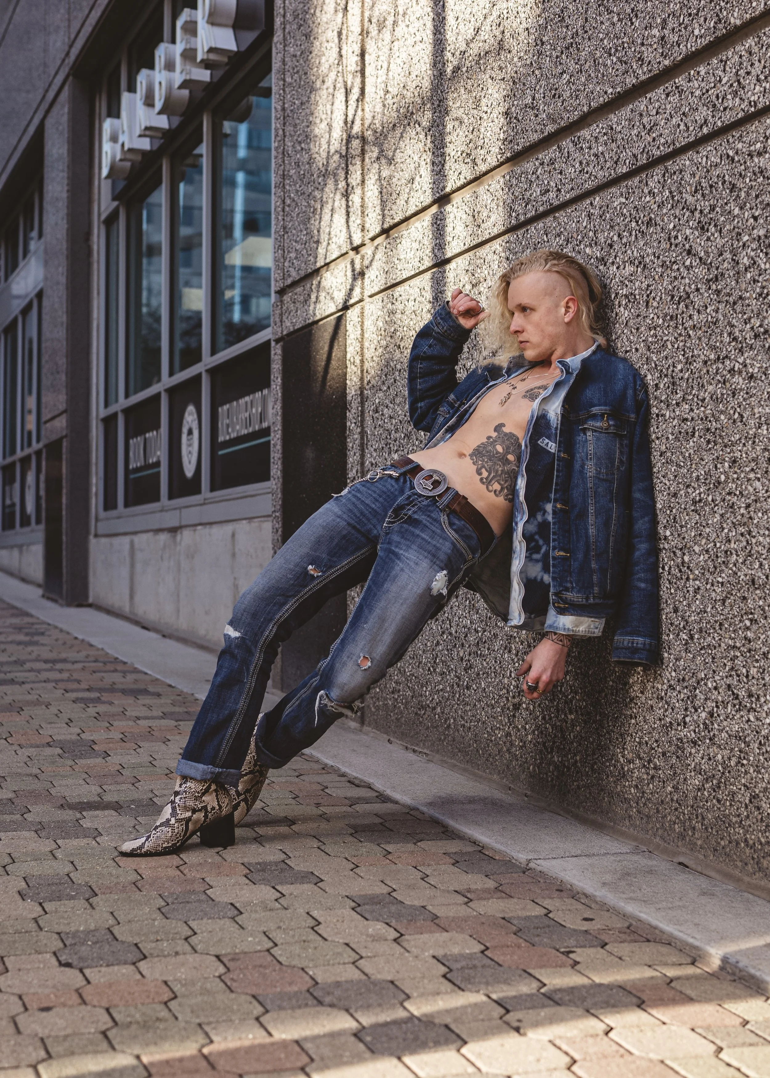 A person with a mohawk hairstyle, wearing a denim jacket, ripped jeans, and snakeskin boots, is leaning against a wall on a city sidewalk.