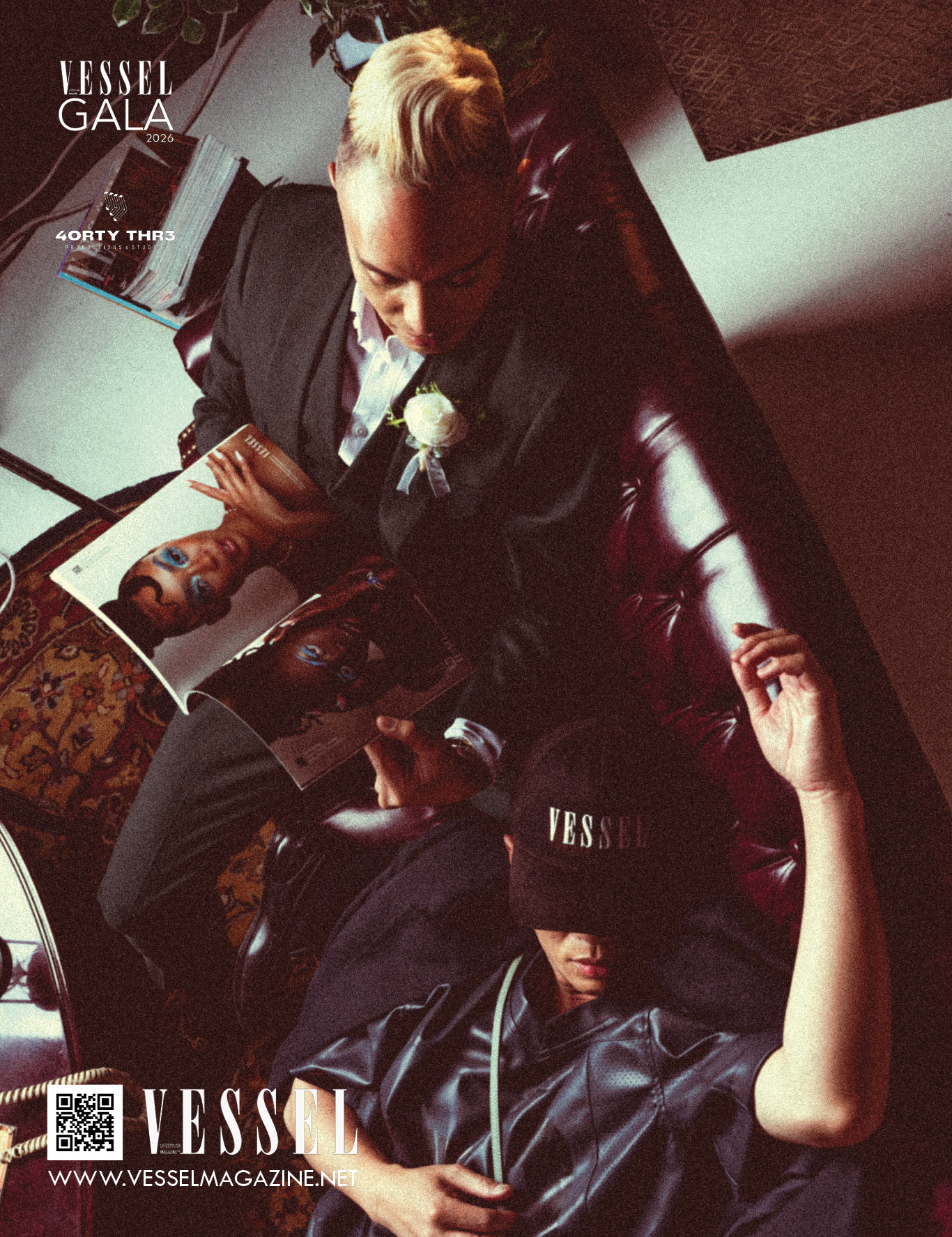 People sitting on a leather sofa, reading magazines, with a magazine featuring a woman with blue eye makeup and a person with platinum blonde hair. The setting appears to be an indoor lounge or waiting area with books and a patterned carpet.