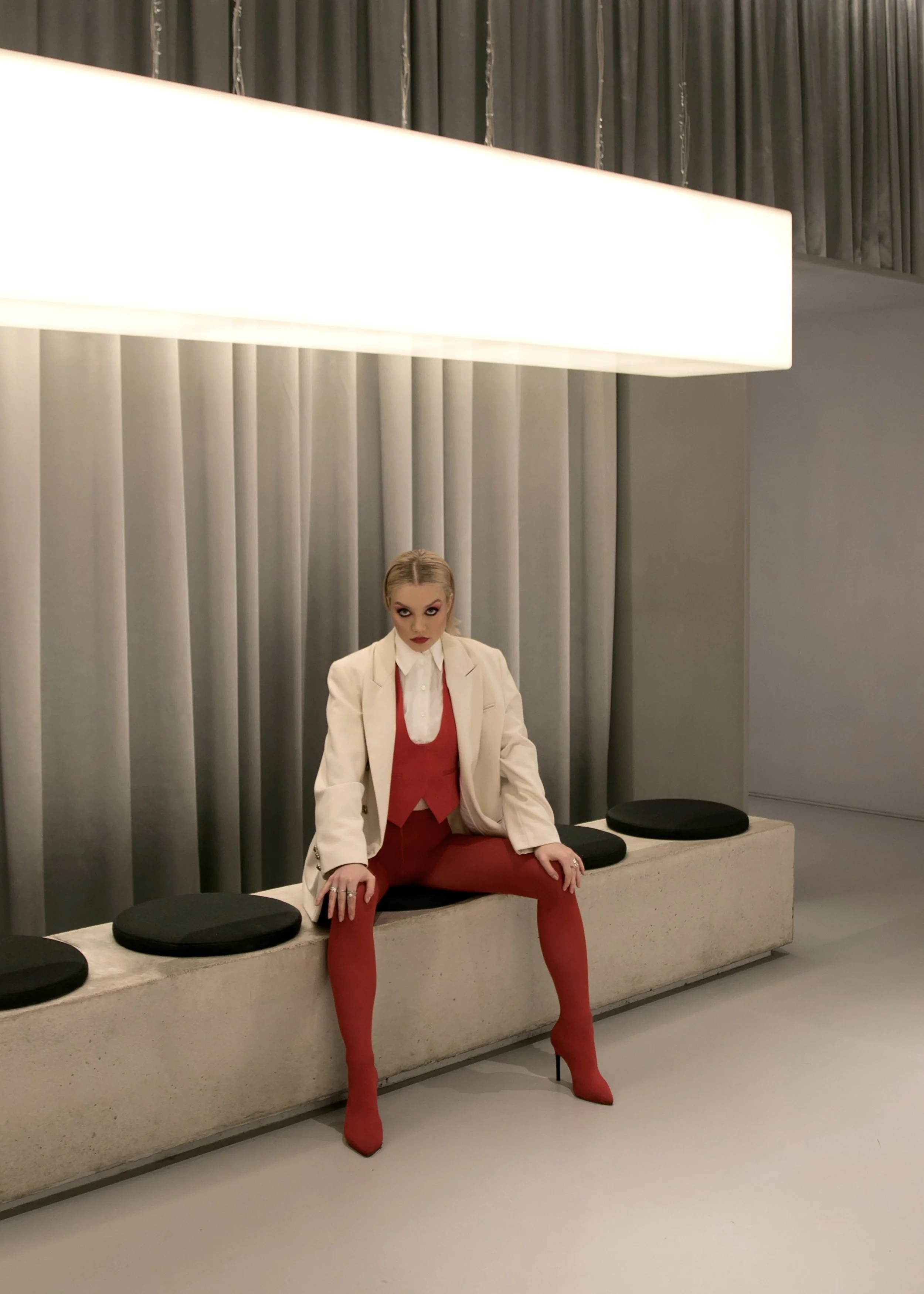 Woman sitting on a concrete bench in front of curtains, wearing a white blazer, red pants, red high heels, and a red vest, with dramatic makeup and blonde hair.