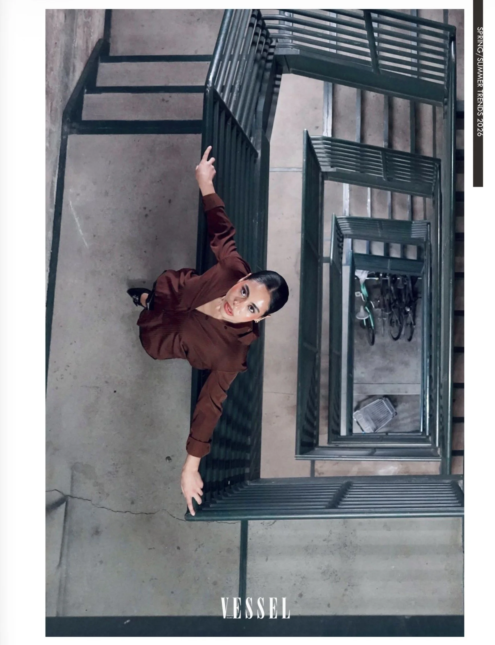 A woman in a brown jacket lying on her side on a concrete floor, reaching out with both hands to touch a large black metal cage or container with multiple compartments. The setting appears to be an industrial or warehouse environment.