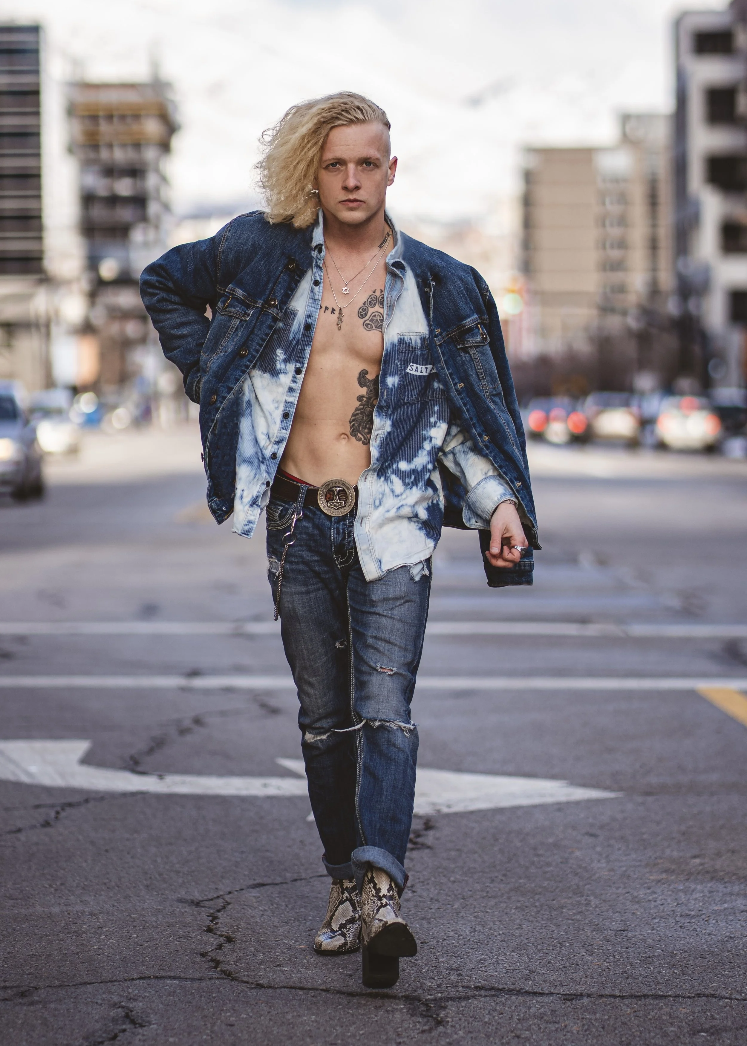 A person walking on a city street, wearing a denim jacket, ripped jeans, snakeskin boots, and various necklaces, with tattoos visible on their chest and neck.