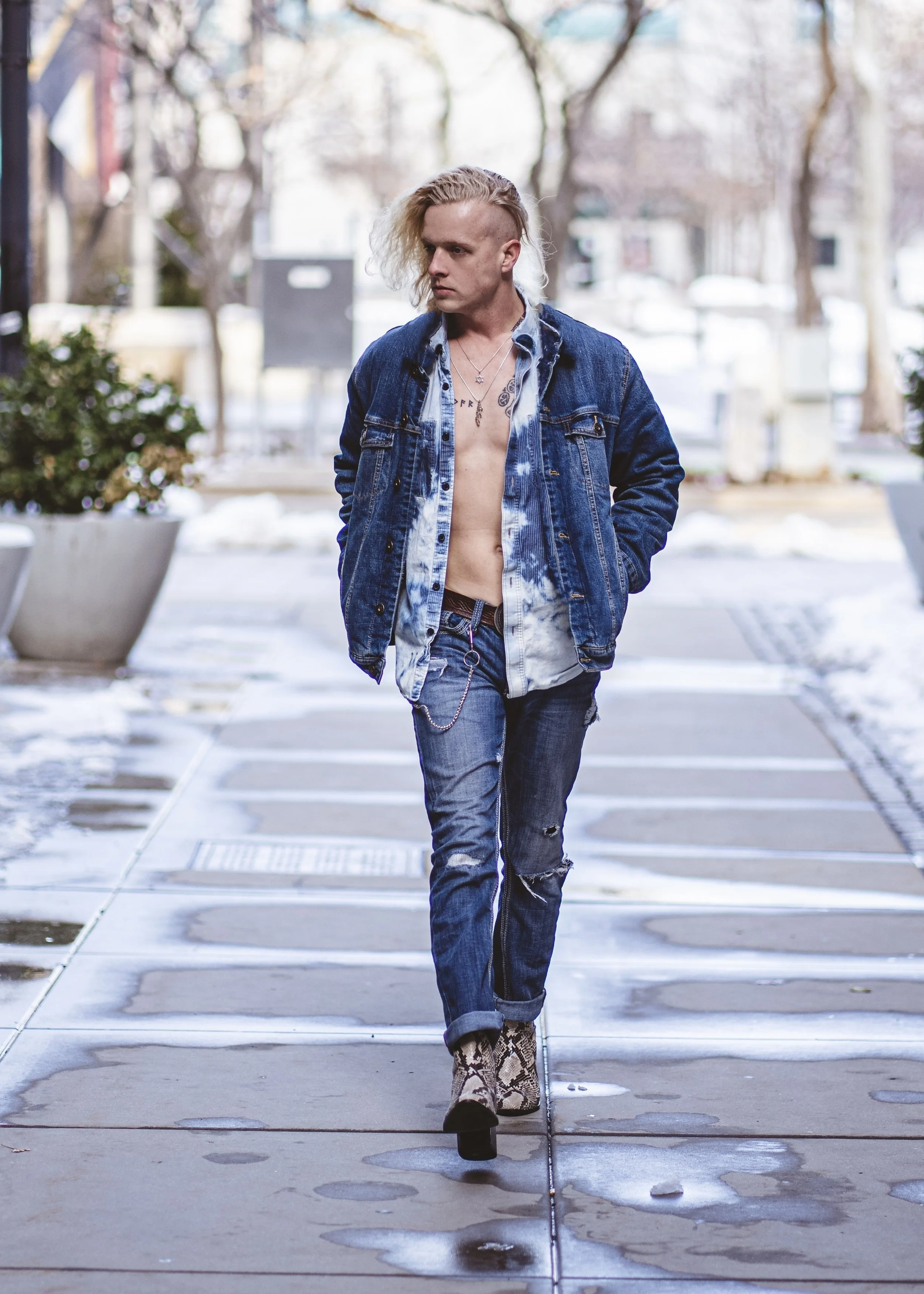 A young man with platinum blond hair, wearing a denim jacket, unbuttoned shirt, ripped jeans, and snakeskin boots, walks on a sidewalk during winter.