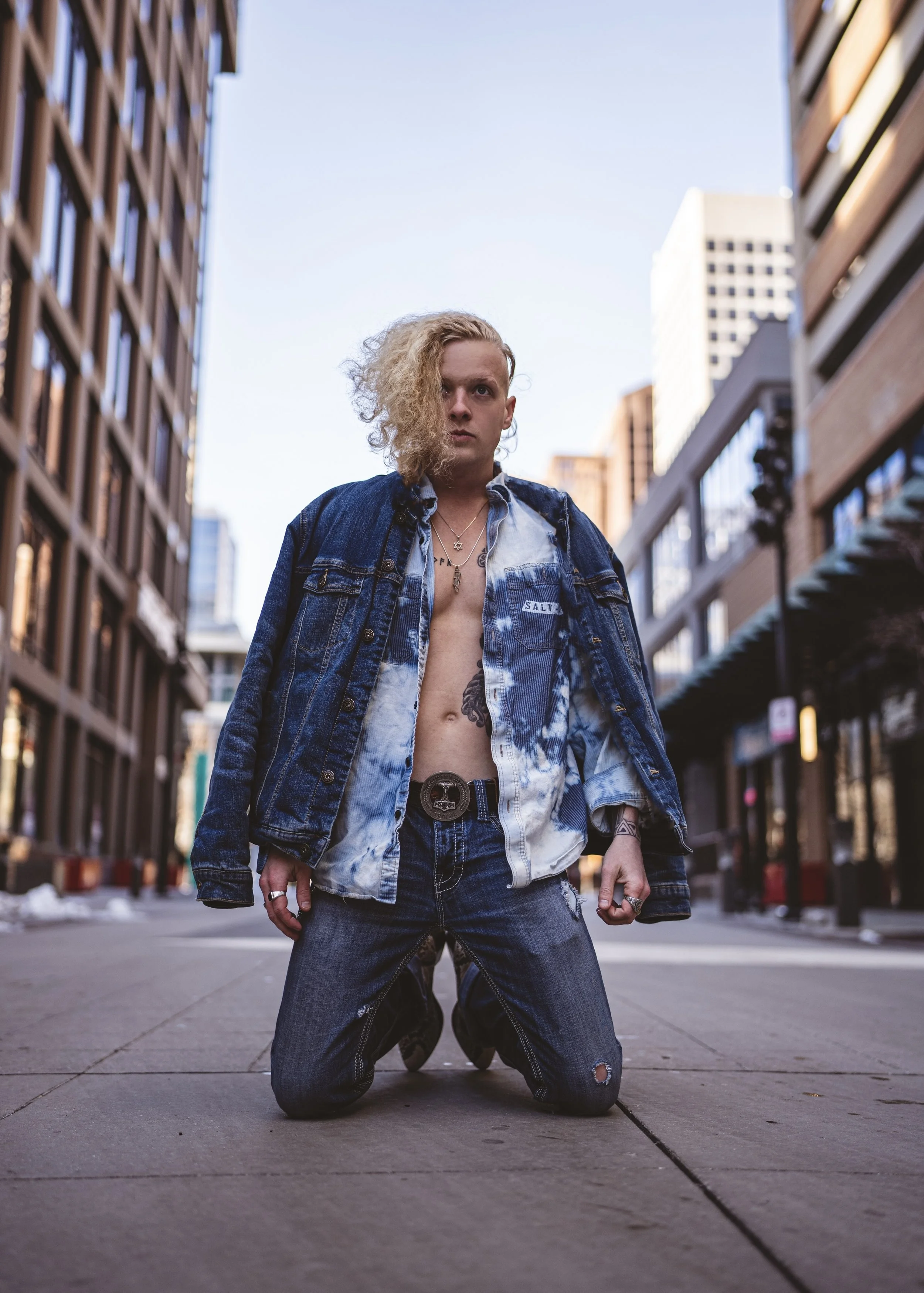 A person with curly blonde hair kneeling on an urban sidewalk, wearing a partially unbuttoned denim jacket and jeans, with tattoos visible on their chest and arm, in a city street with tall buildings on both sides.