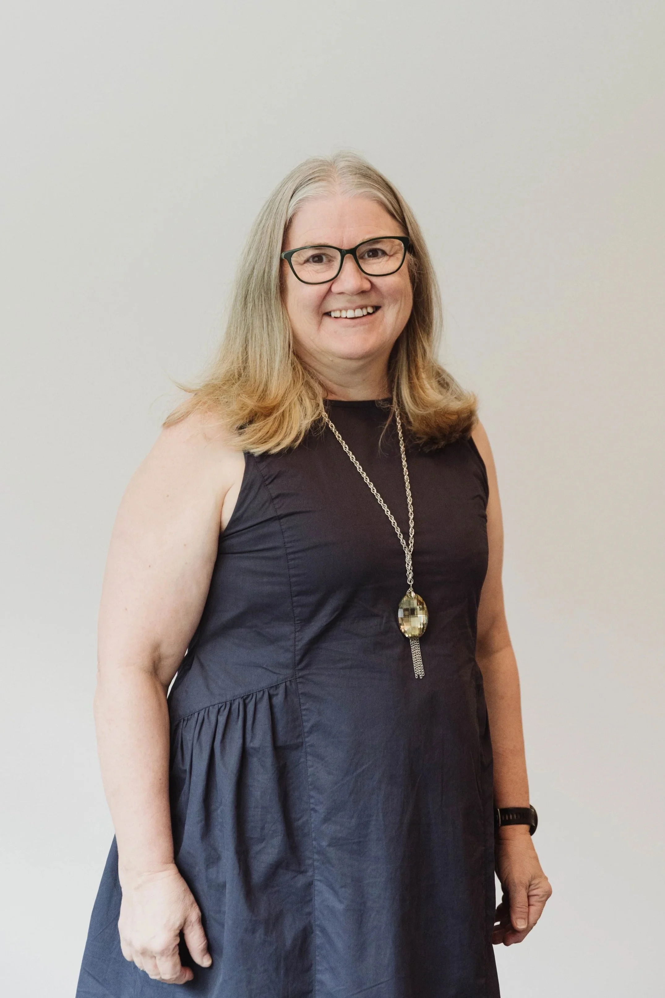A woman with shoulder-length blonde hair, glasses, and a black sleeveless dress standing against a plain light-colored wall, smiling at the camera.