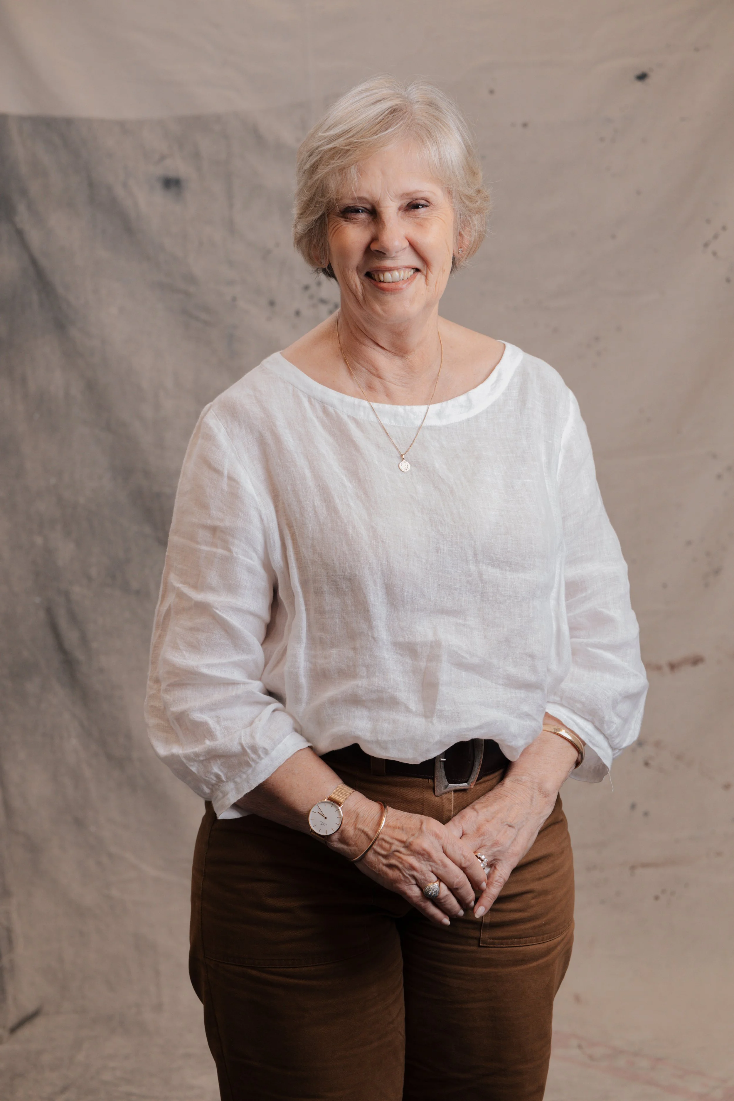 An elderly woman with short brown hair smiling, wearing a striped button-up shirt, white pants, and gold jewelry, standing in front of a plain backdrop.