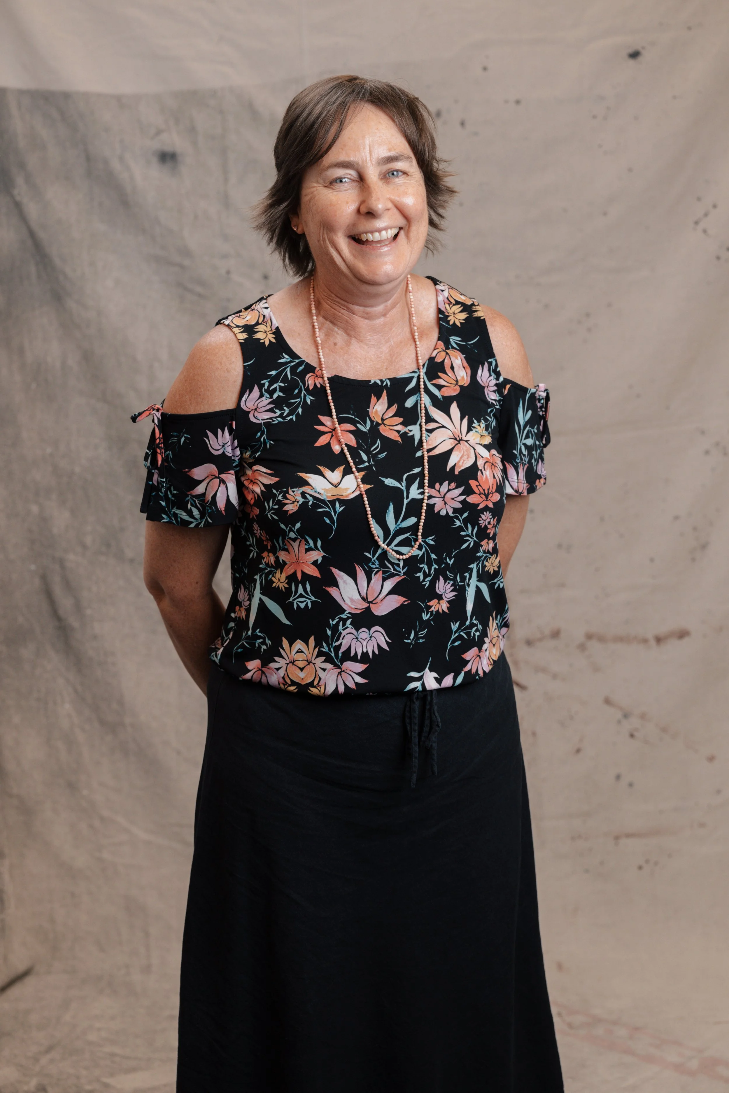 A woman with brown hair, smiling, wearing a colorful blouse, black cardigan, and jeans, standing in front of a beige backdrop.