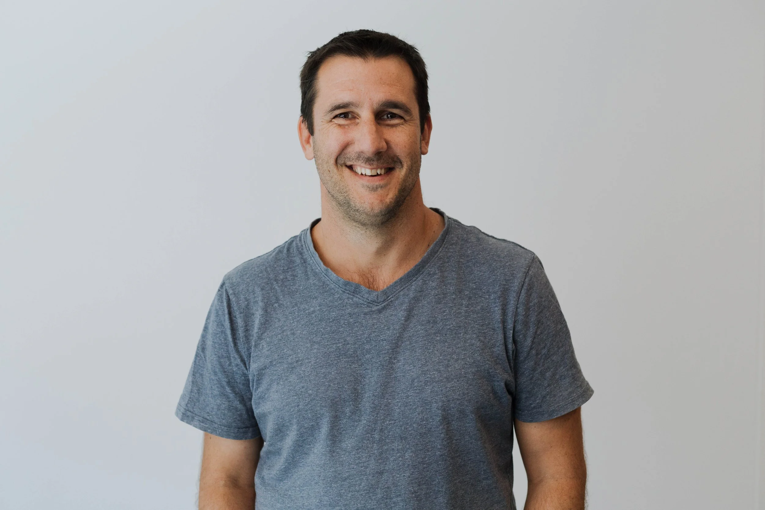 A smiling man with dark hair and stubble, wearing a grey T-shirt, standing against a plain white background.