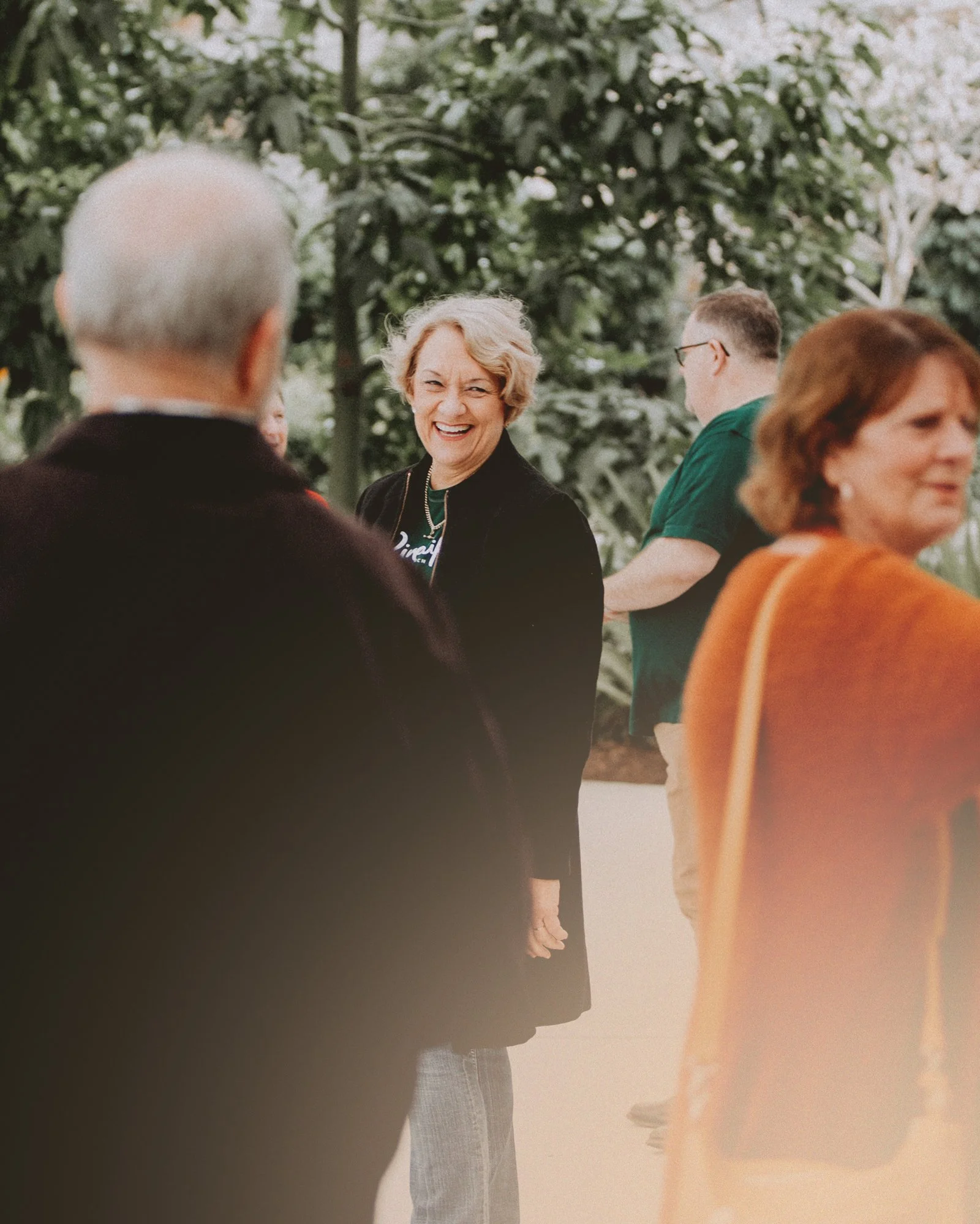 A woman with short blonde hair smiling and laughing at a social gathering outdoors, surrounded by other people, with greenery in the background.
