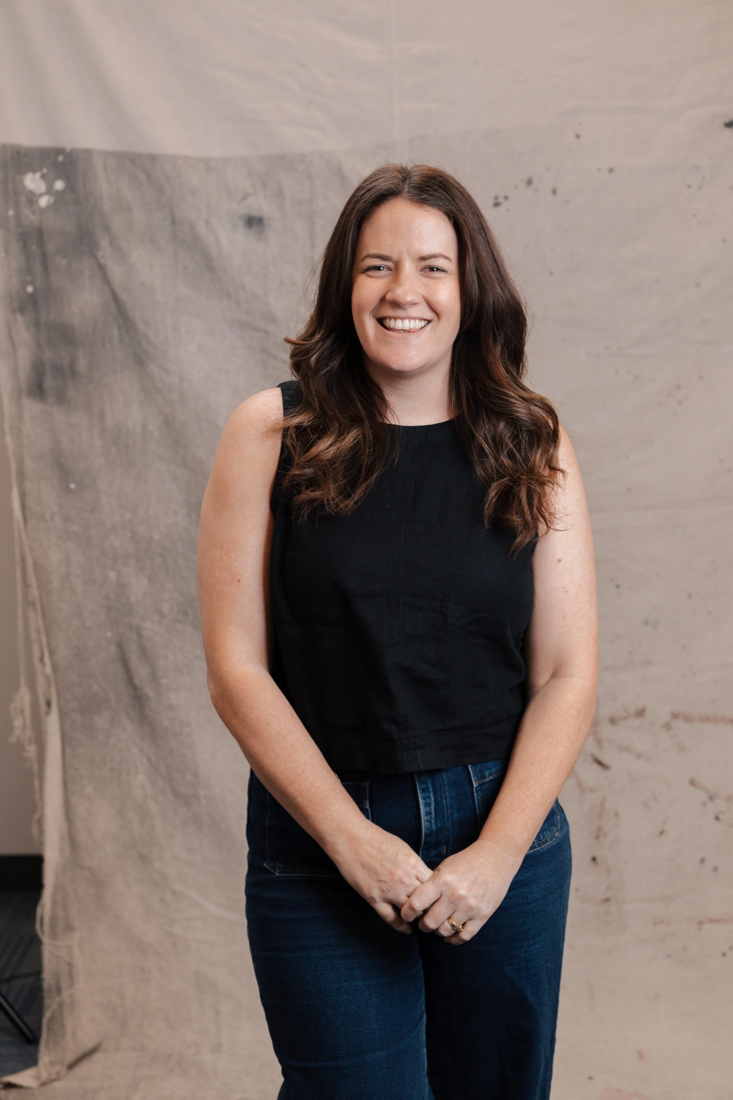 A woman with shoulder-length wavy brown hair, smiling, wearing a green sweater and blue jeans, standing against a beige fabric backdrop.