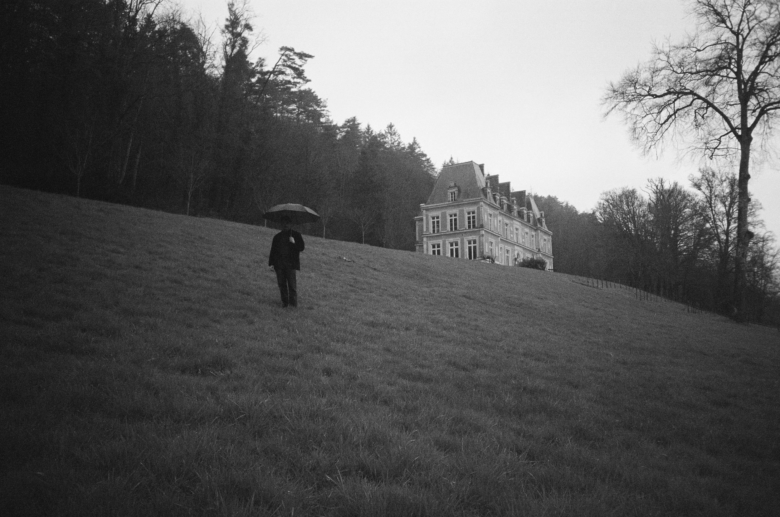 A person holding an umbrella walking on a grassy hill in front of a large, ornate house surrounded by trees, with an overcast sky.