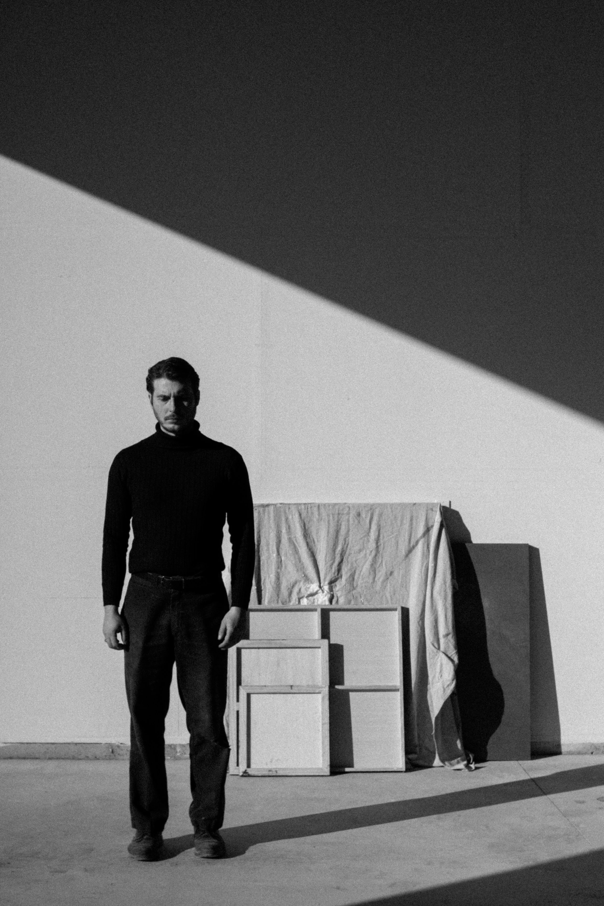 A black and white photo of a man in a black turtleneck and jeans standing in front of a simple indoor backdrop, with one person crouching next to a then-covered table and a person lying on the ground. The setting appears to be a minimalist or abstract space, with a shadow cast on the floor.