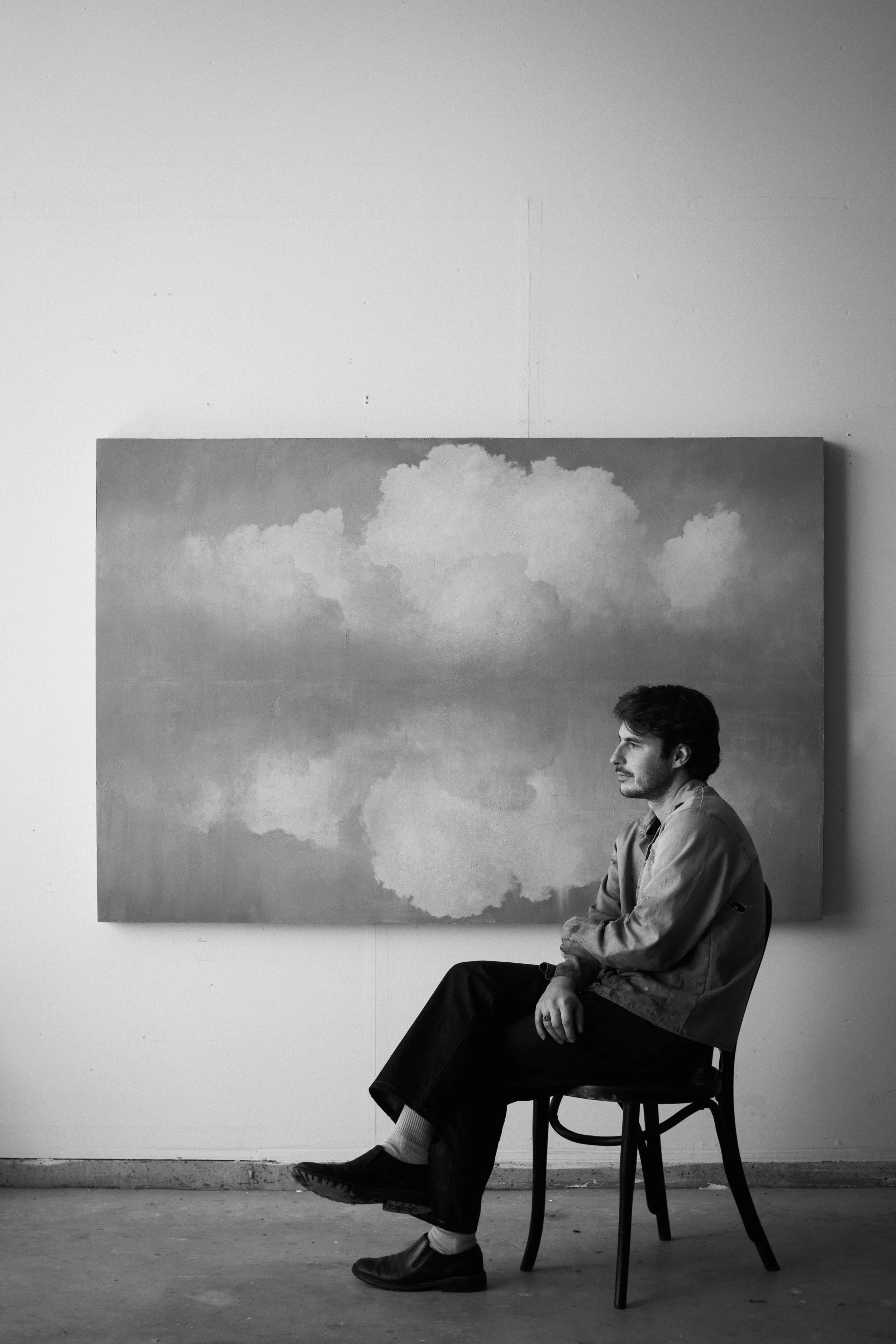 A man with dark hair, wearing a light-colored jacket, sitting on a wooden chair with his right arm resting on his lap, looking to the right, in an art gallery with a monochrome painting of clouds on the wall behind him.