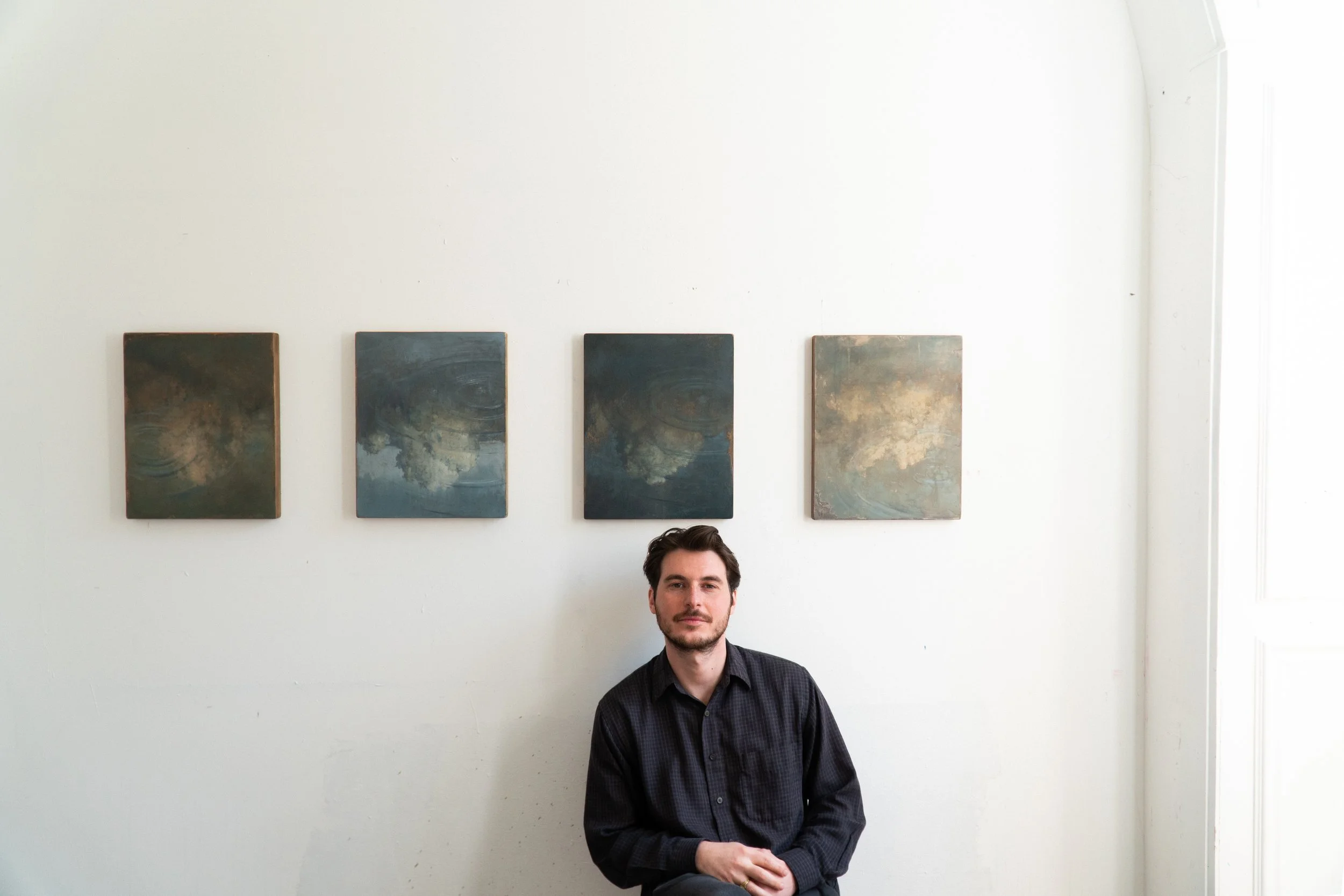 A man sits in front of a white wall with four abstract paintings hanging above him. The man has dark hair and a beard, and is wearing a black, plaid button-up shirt.