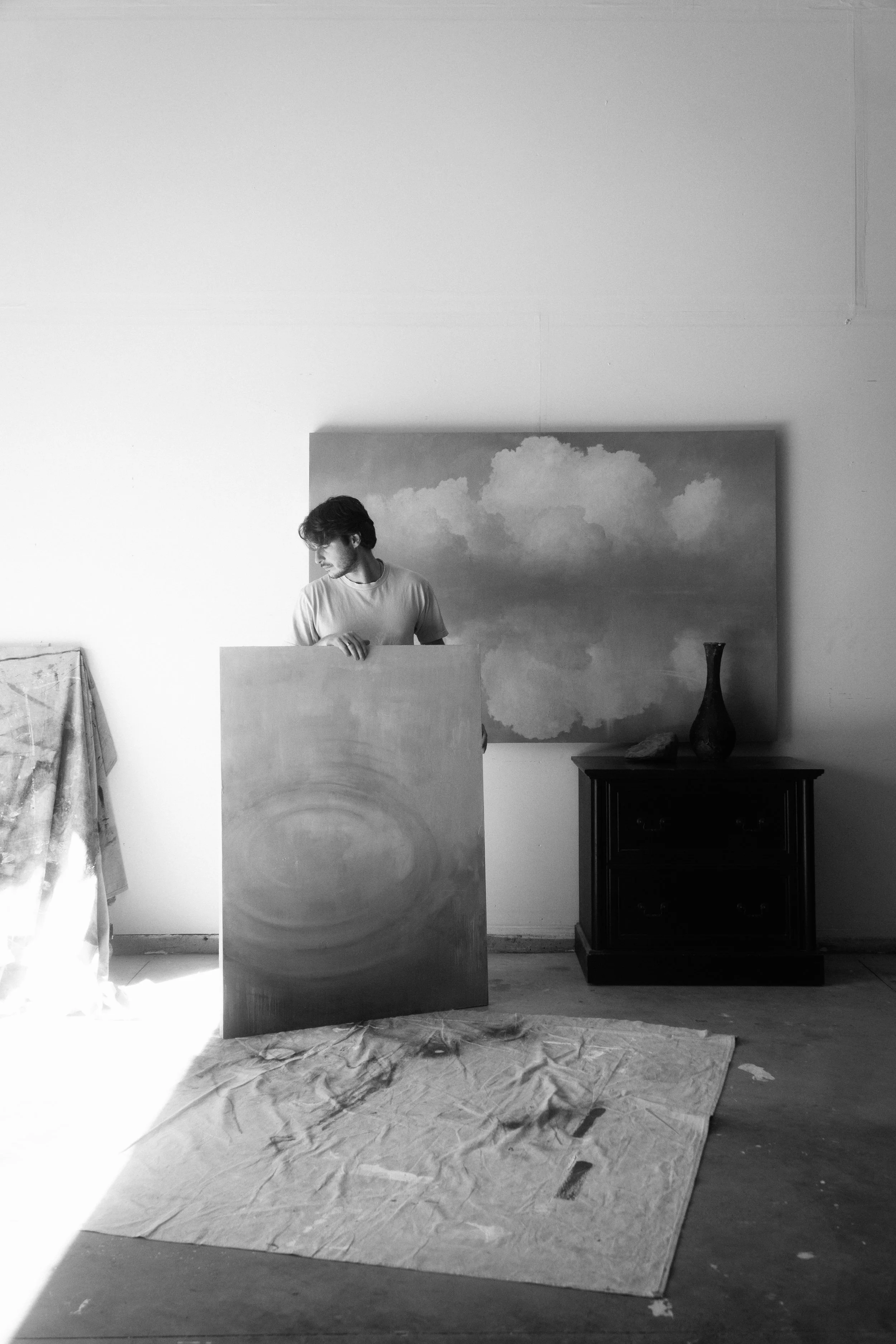 A man with dark hair and a beard looking at a large abstract painting with a swirling circular design in a room with white walls and wooden furniture.