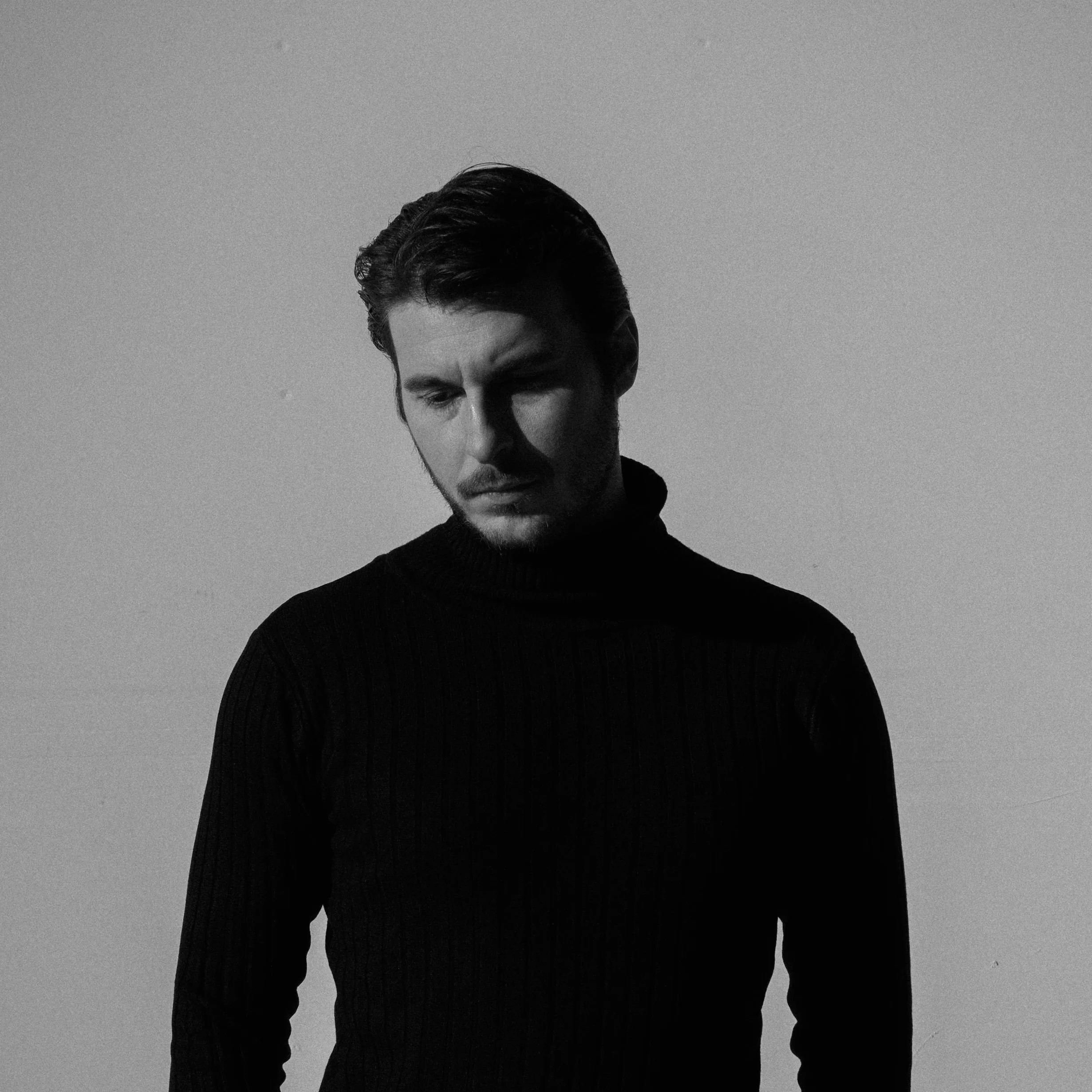 A black and white portrait of a man with dark, slicked-back hair, wearing a dark turtleneck sweater, looking downward, against a plain gray background.