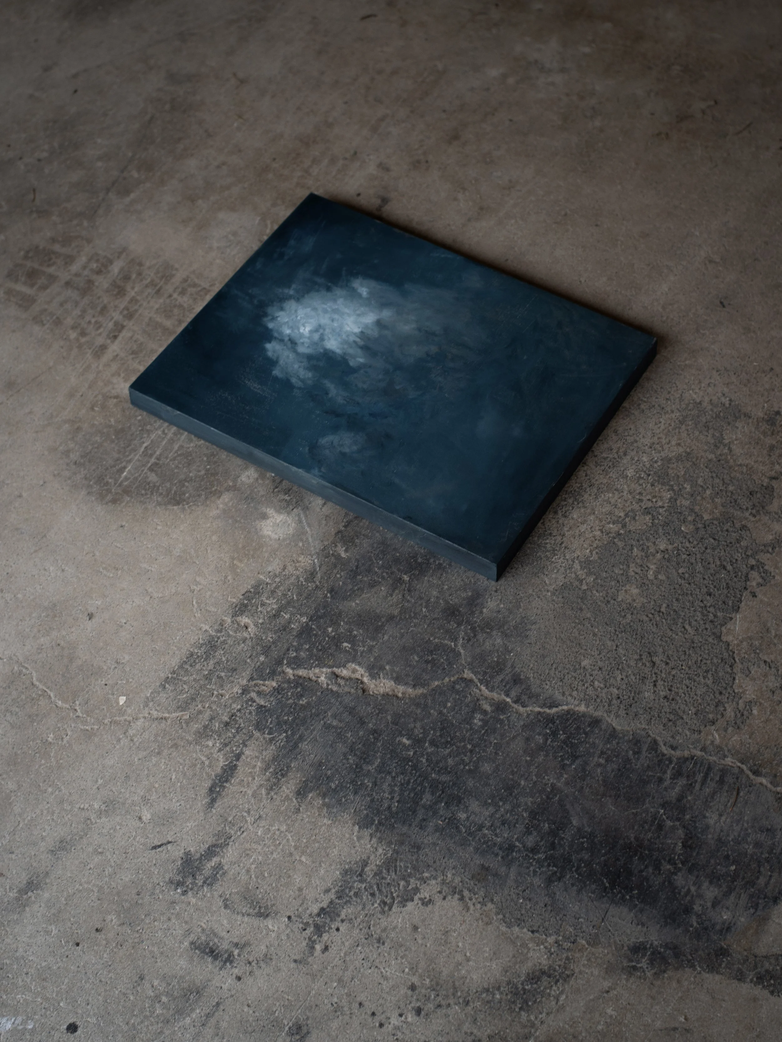 A square painting with abstract blue and white cloud-like patterns resting on a worn concrete floor.