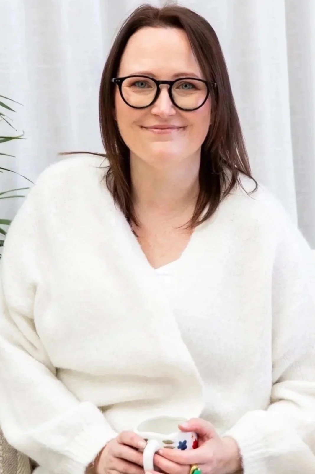 A woman with shoulder-length brown hair, glasses, and a white sweater smiling while holding a white mug with a flower pattern, sitting in front of a white curtain and a green plant.