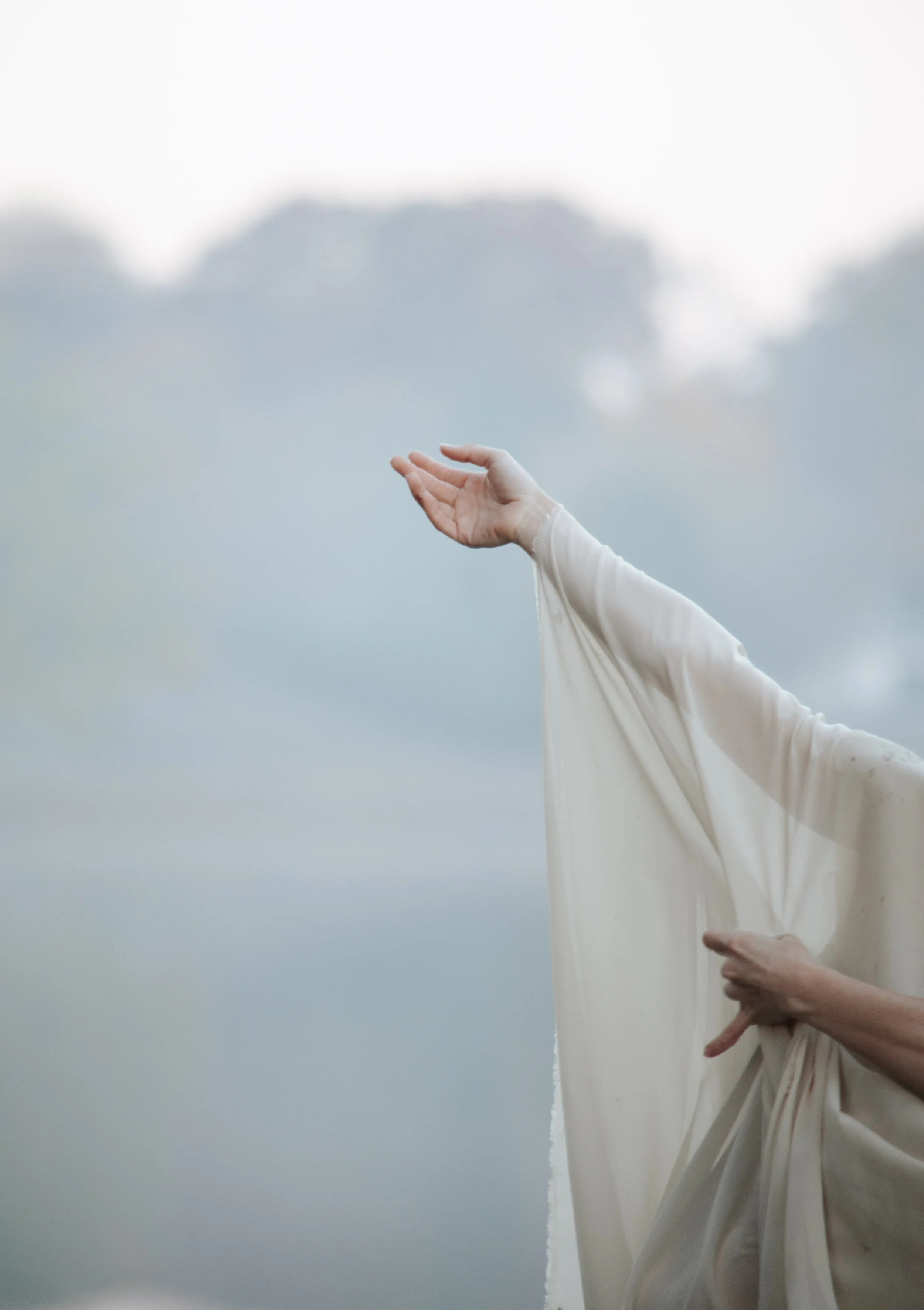 Close-up of a person's hand and arm with a sheer fabric draped over the arm extending outward against a blurry outdoor background.