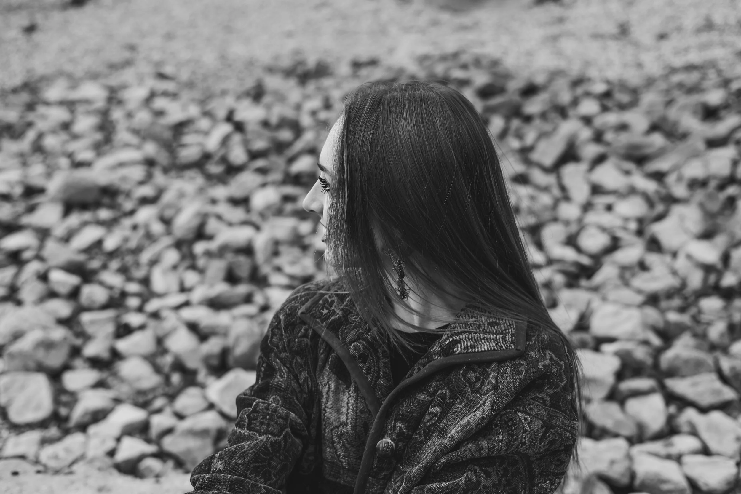 A black and white image of a young woman wearing a coat, smiling, and looking into the distance with rocks in the background