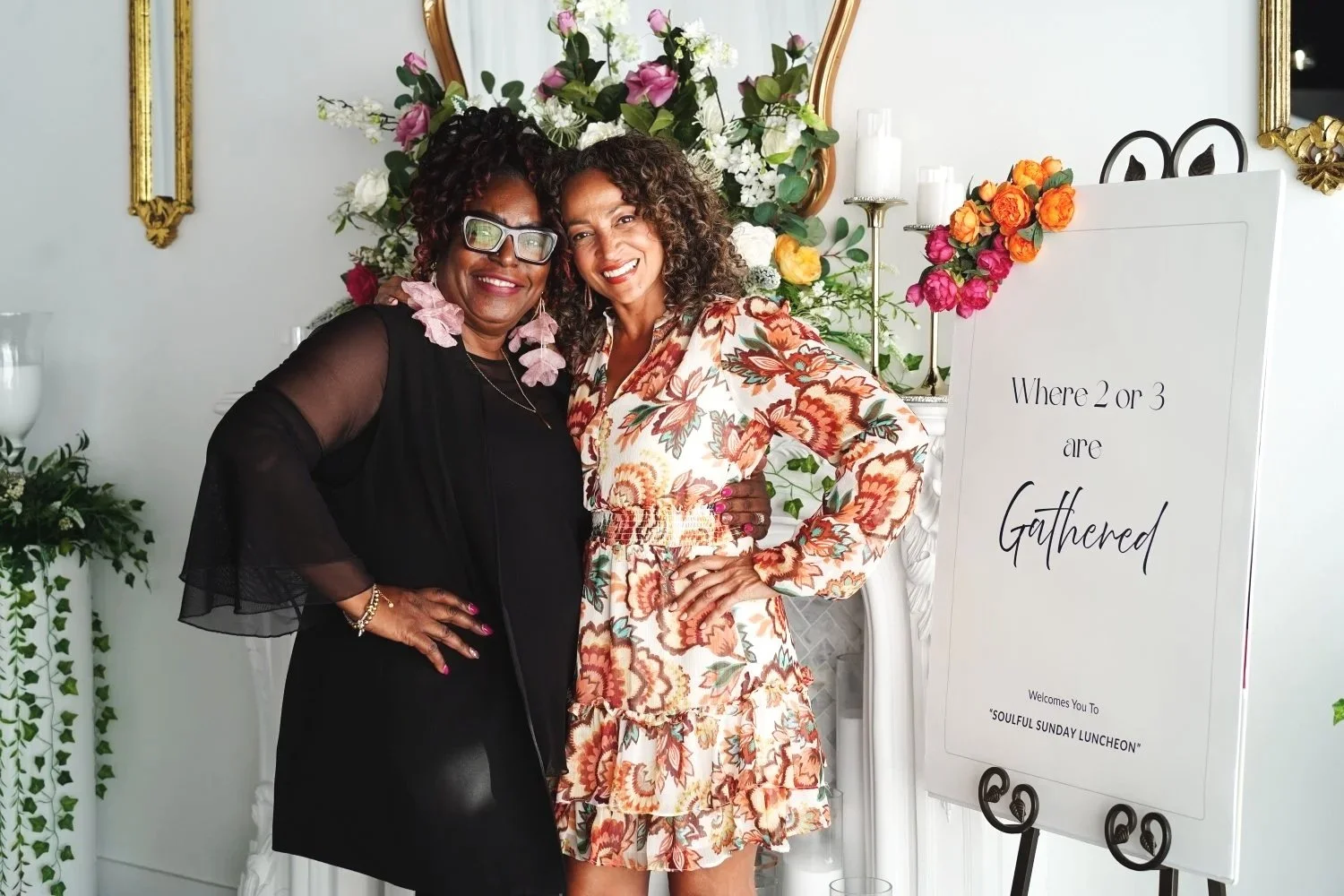 Two women standing together, smiling, at a floral-themed event with a sign that reads 'Where 2 or 3 are Gathered' and decorated with flowers in the background.