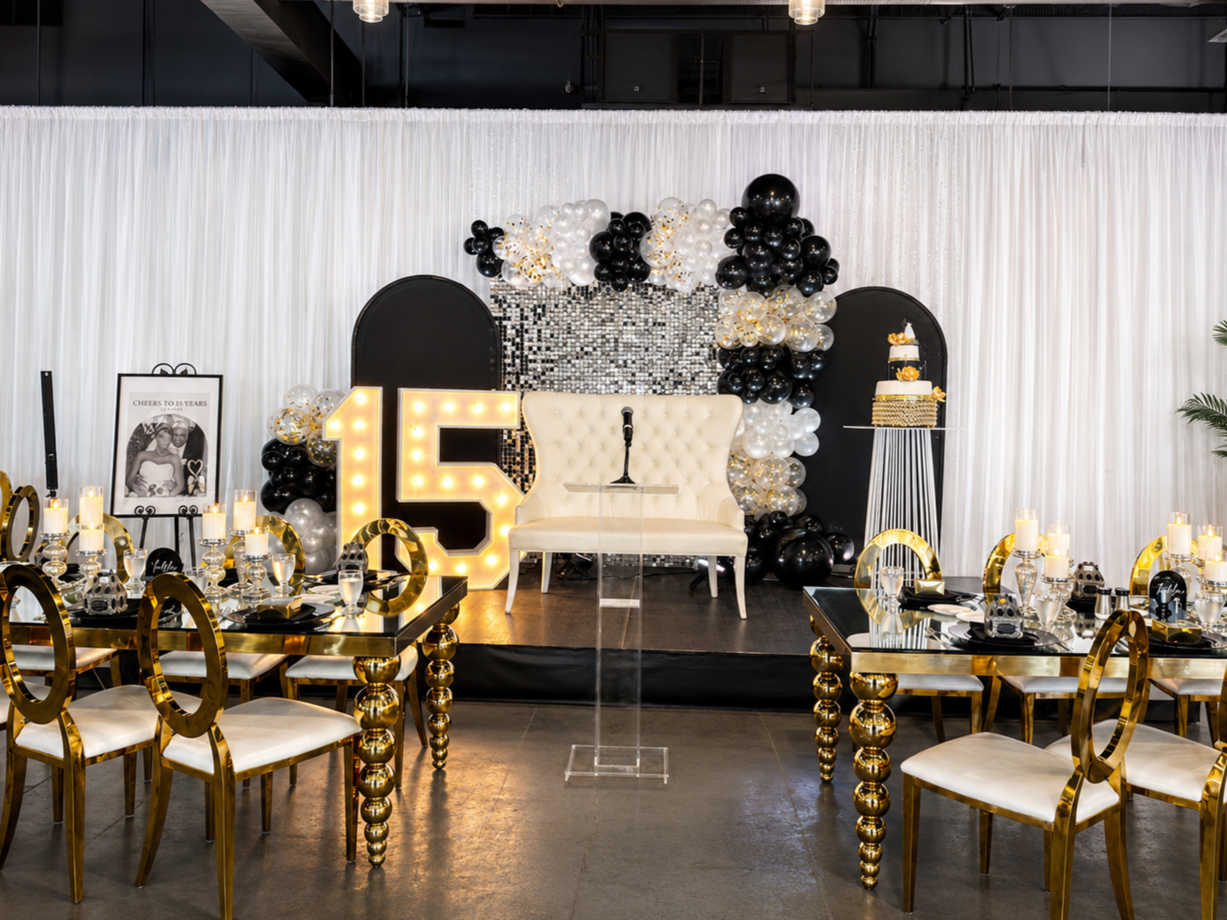 Elegant celebration setup with a backdrop of white curtains, black and gold balloons, and a mosaic tile wall. A large illuminated number 15 is lit up next to a white tufted couch with a microphone in front. Tables are decorated with gold and white ac