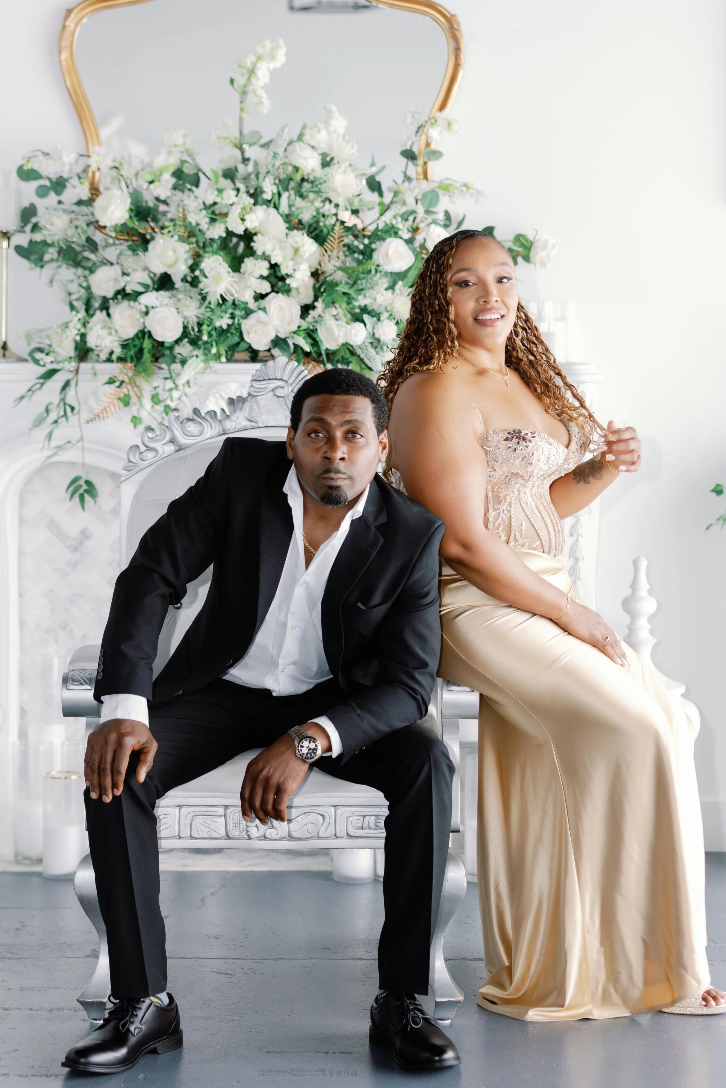 A man and a woman sitting on a white ornate bench with a large floral arrangement behind them. The man is wearing a black suit, white shirt, and black shoes, leaning forward with one hand on his knee. The woman is in a strapless beige gown, sitting w