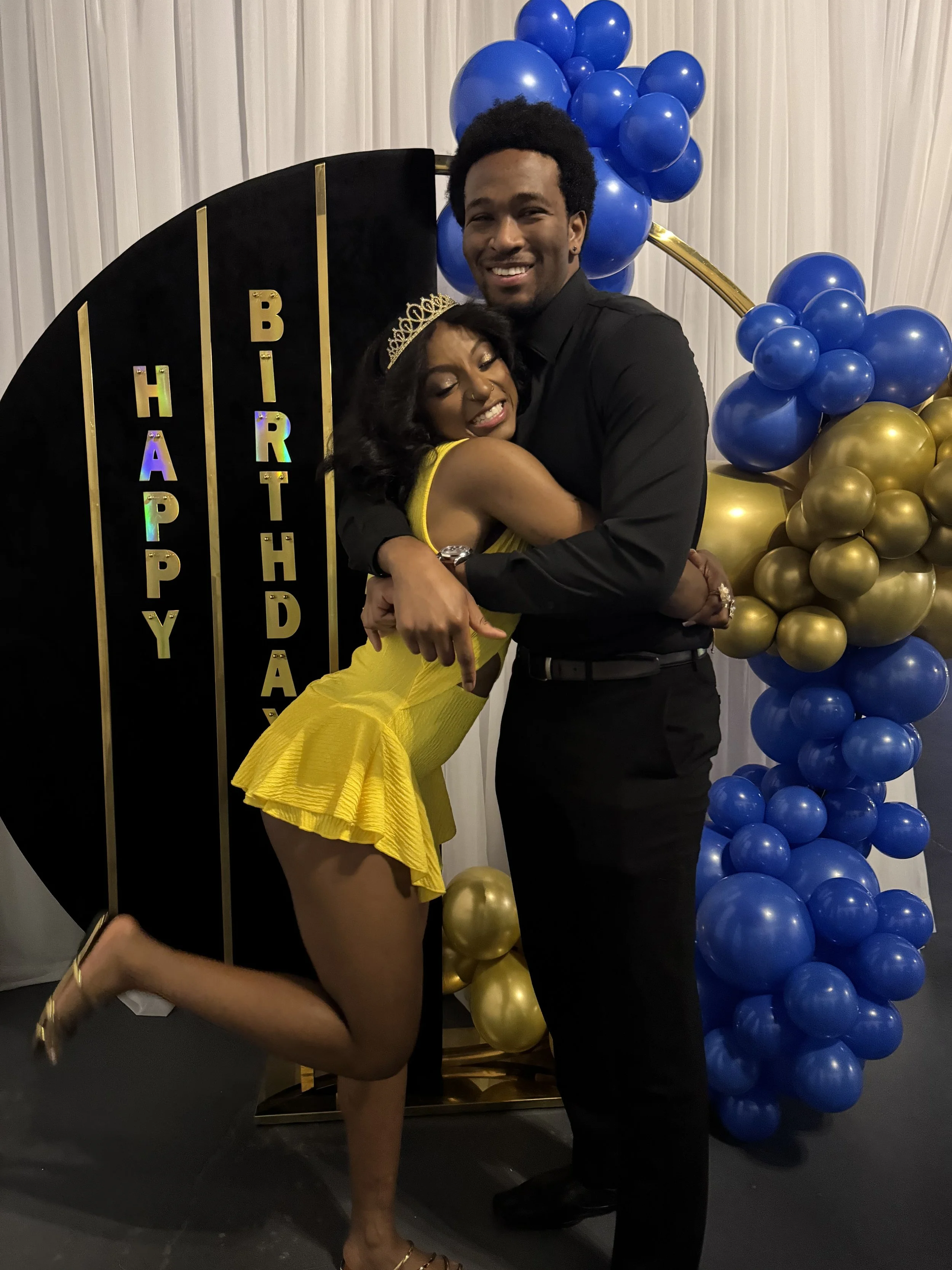A woman and a man are hugging in front of a decorative background with blue and gold balloons. The woman is wearing a yellow dress and a tiara, and the man is in a black shirt and pants, smiling at the camera.