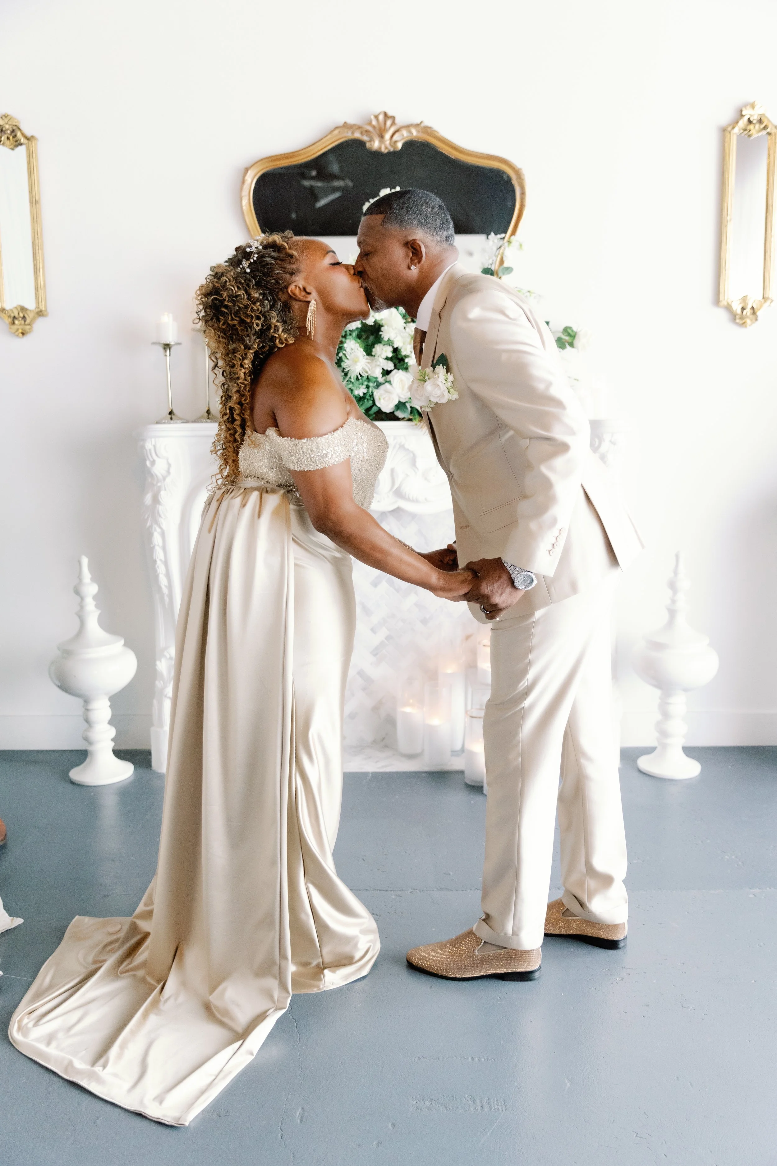 A bride and groom sharing a kiss at their wedding ceremony, with elegant vintage-style decor and a white fireplace with floral arrangements behind them.