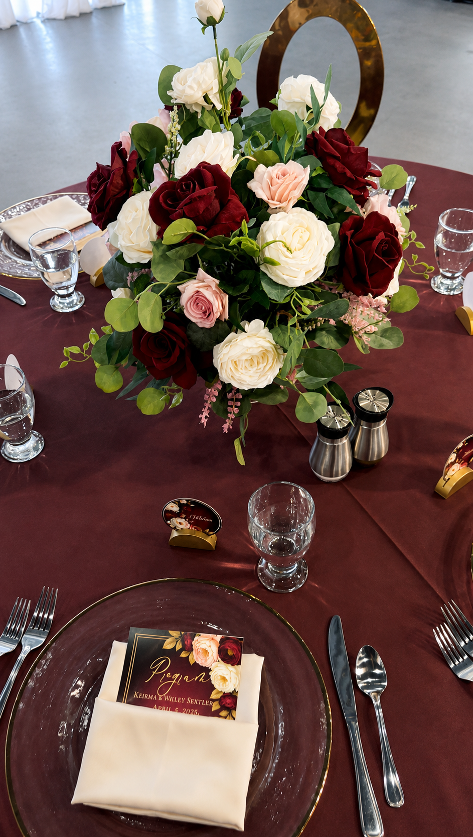 A formal table setting with a dark red tablecloth, a large floral centerpiece with white, red, and blush pink roses, and a gold ring flower. There are clear glass water goblets, stainless steel salt and pepper shakers, gold place card holders, a menu