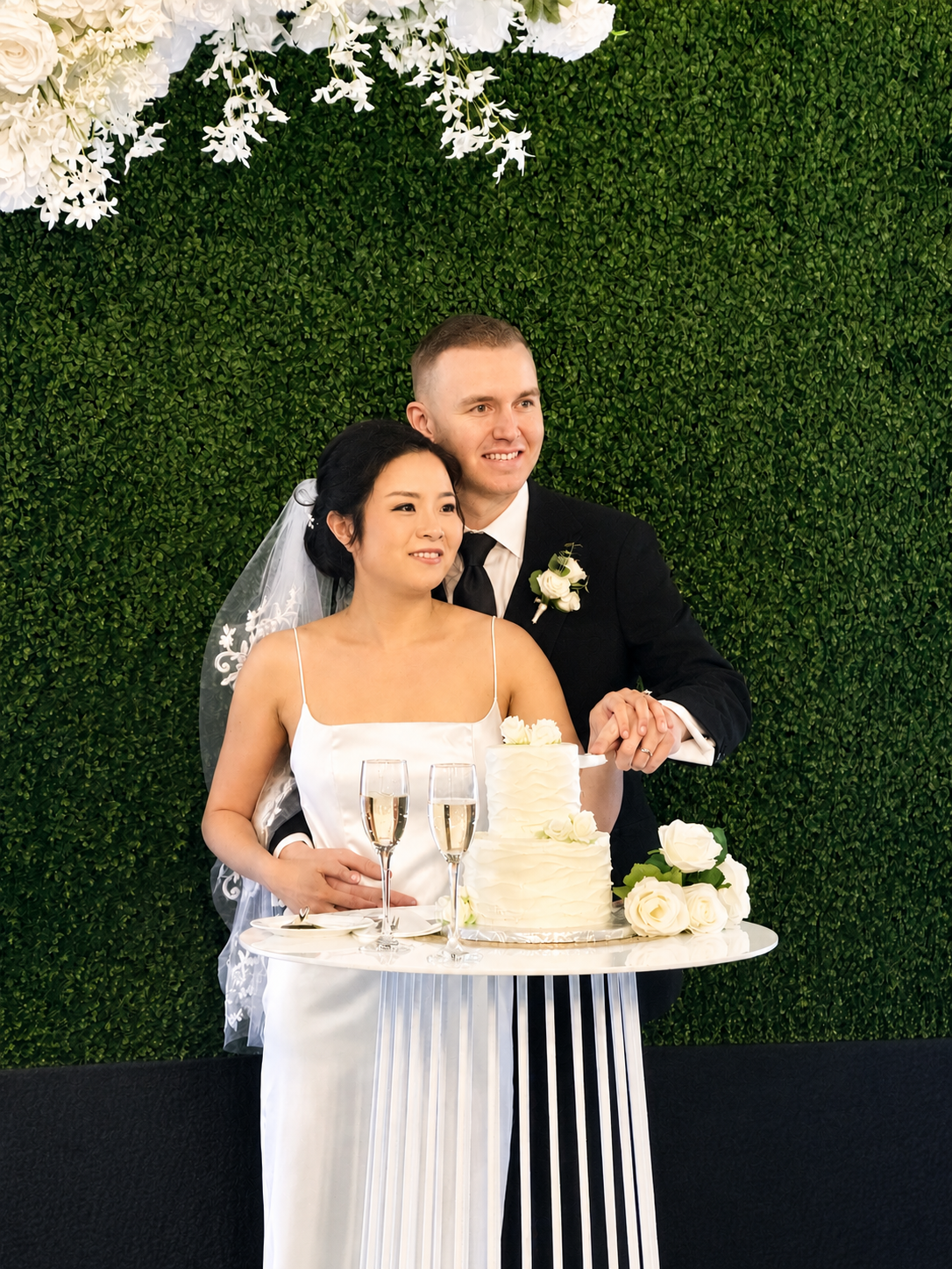 A bride and groom at their wedding reception, cutting a white wedding cake with white roses, standing in front of a green leafy backdrop decorated with white flowers, two glasses of champagne on the table.