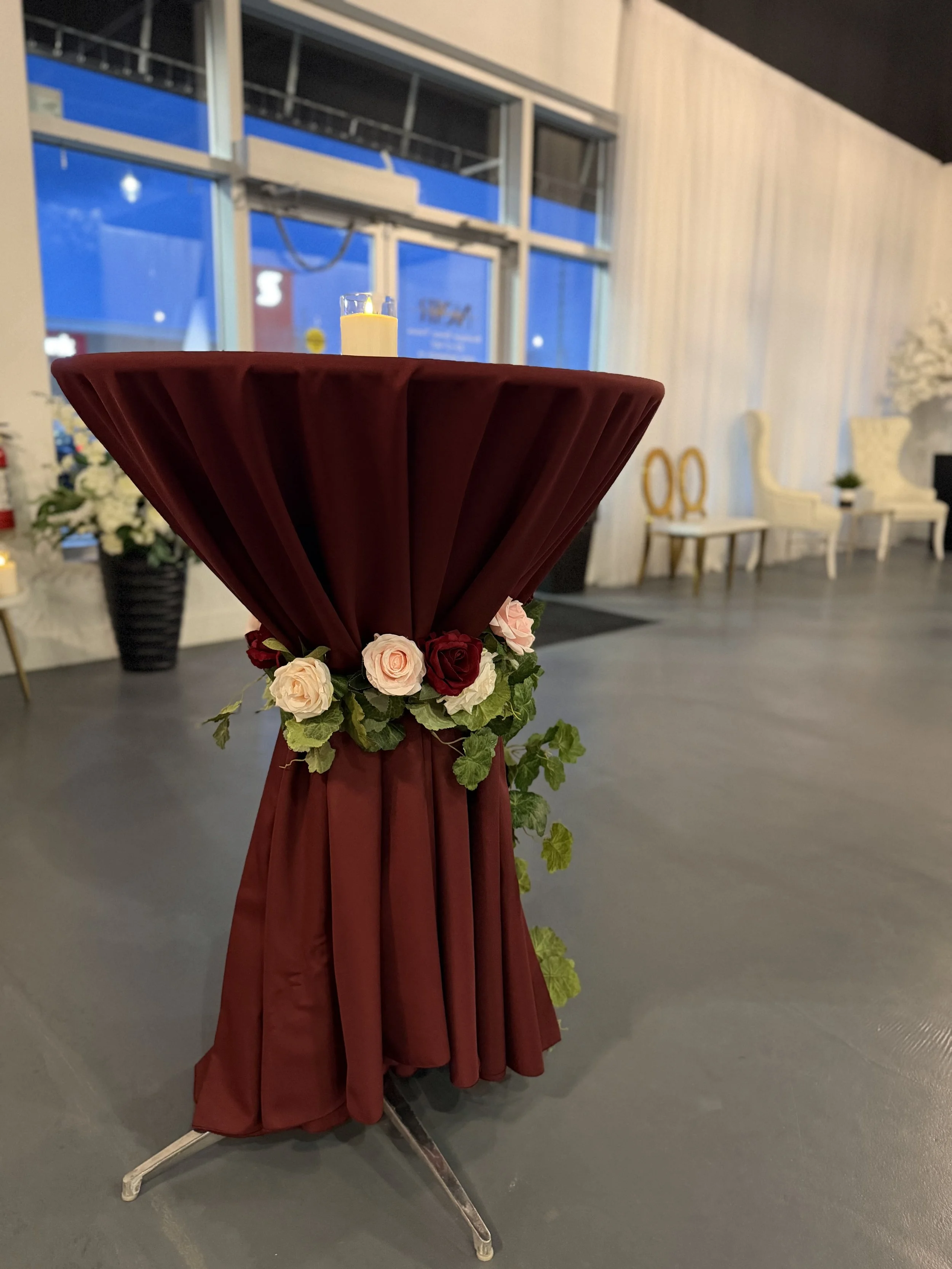 A round table draped with a maroon tablecloth decorated with pink and red roses and green leaves, with a small lit candle on top, in a well-lit indoor setting with white chairs and floral arrangements in the background.