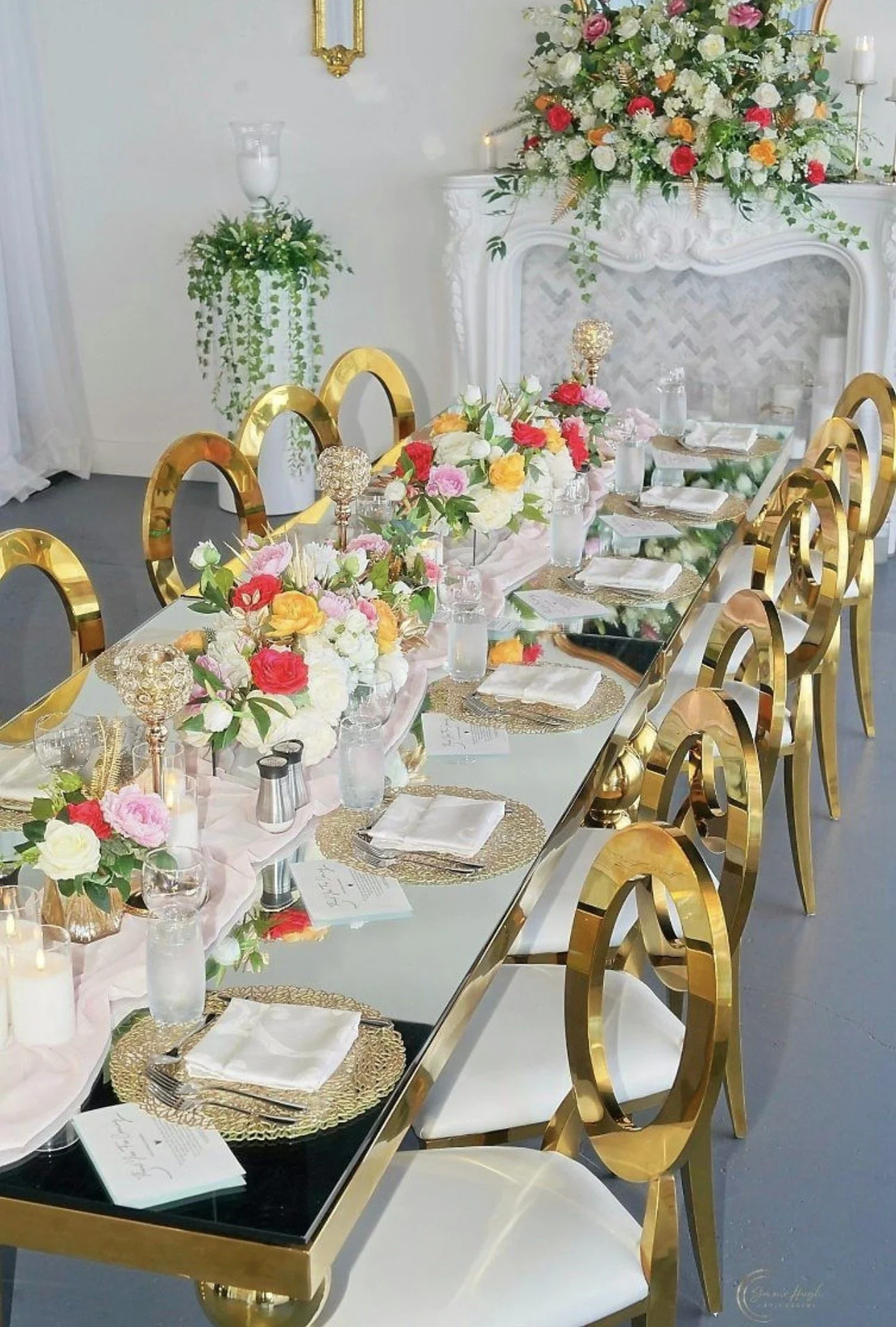 Elegant dining table set for a wedding with gold chairs, floral centerpieces, candles, and place settings with white napkins and gold chargers, with a large floral arrangement on a white ornate fireplace in the background.