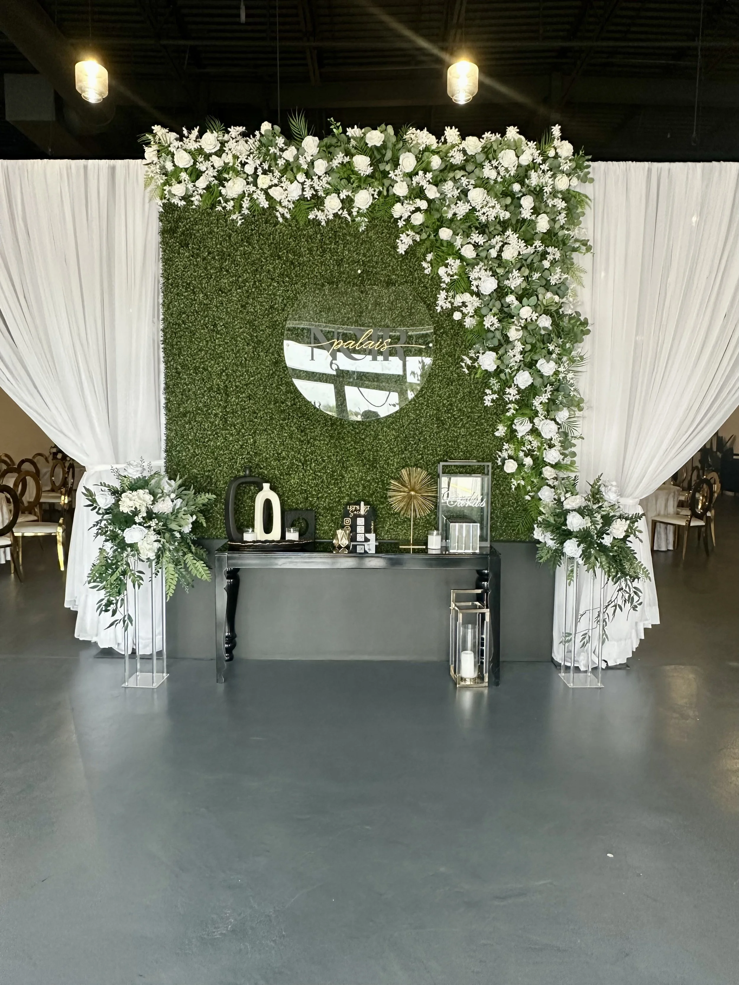 Elegant wedding or event decor with a green leafy backdrop adorned with white flowers and a mirror with gold lettering that reads 'Palais.' There are white drapes on either side, floral arrangements, and decorative items including candles and modern 