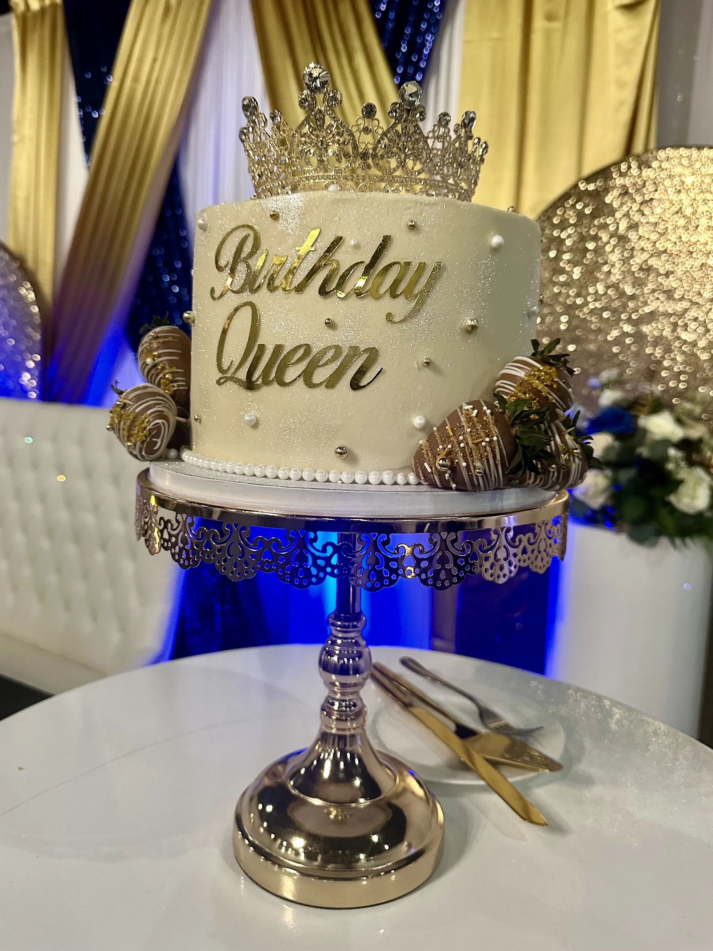 A birthday cake with a gold crown on top, decorated with pearls and gold beads, with the words 'Birthday Queen' in gold lettering, displayed on a silver stand with a white table and gold utensils nearby.